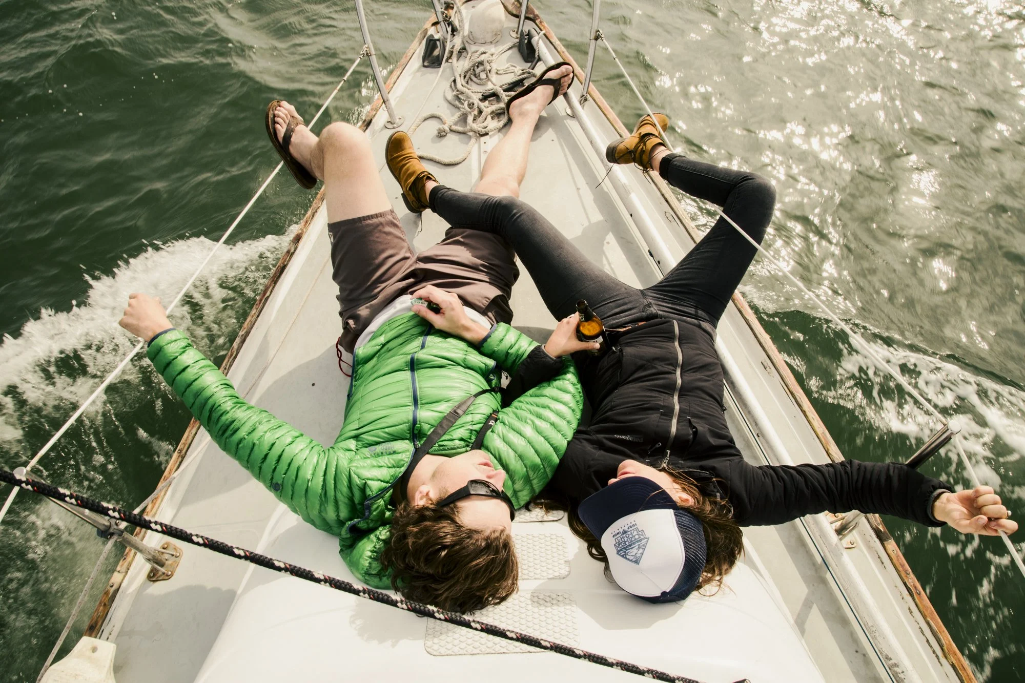 Three people lying on a sailboat deck: one person with a green jacket and sunglasses, another in black leggings and boots, and the third in brown shorts and flip-flops. They are relaxing on the boat with water surrounding them, and one person is hold