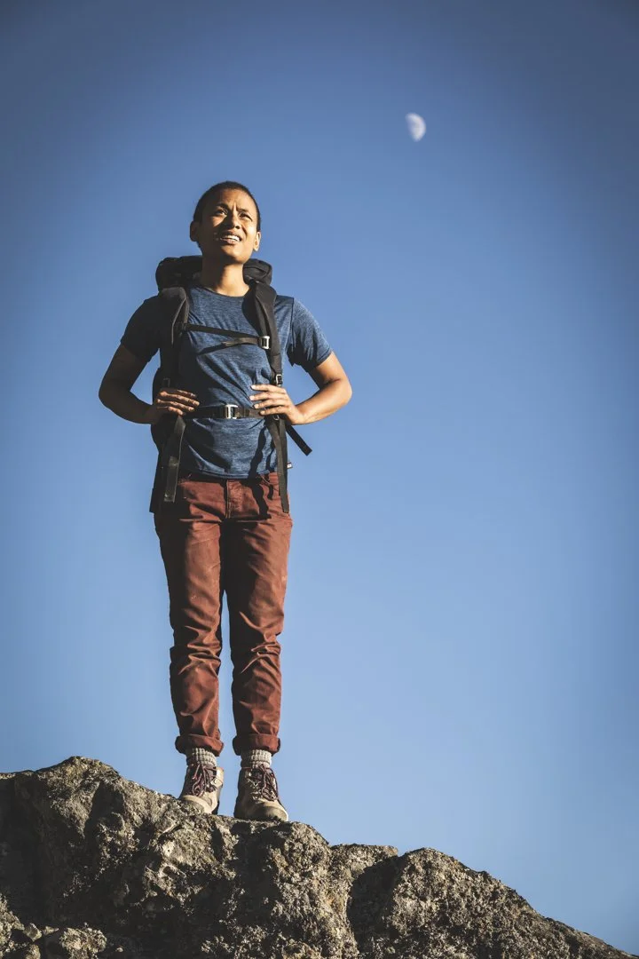 Woman with backpack standing on rocks outdoors looking into distance with moon in sky, photographed by Fieldborn Creative