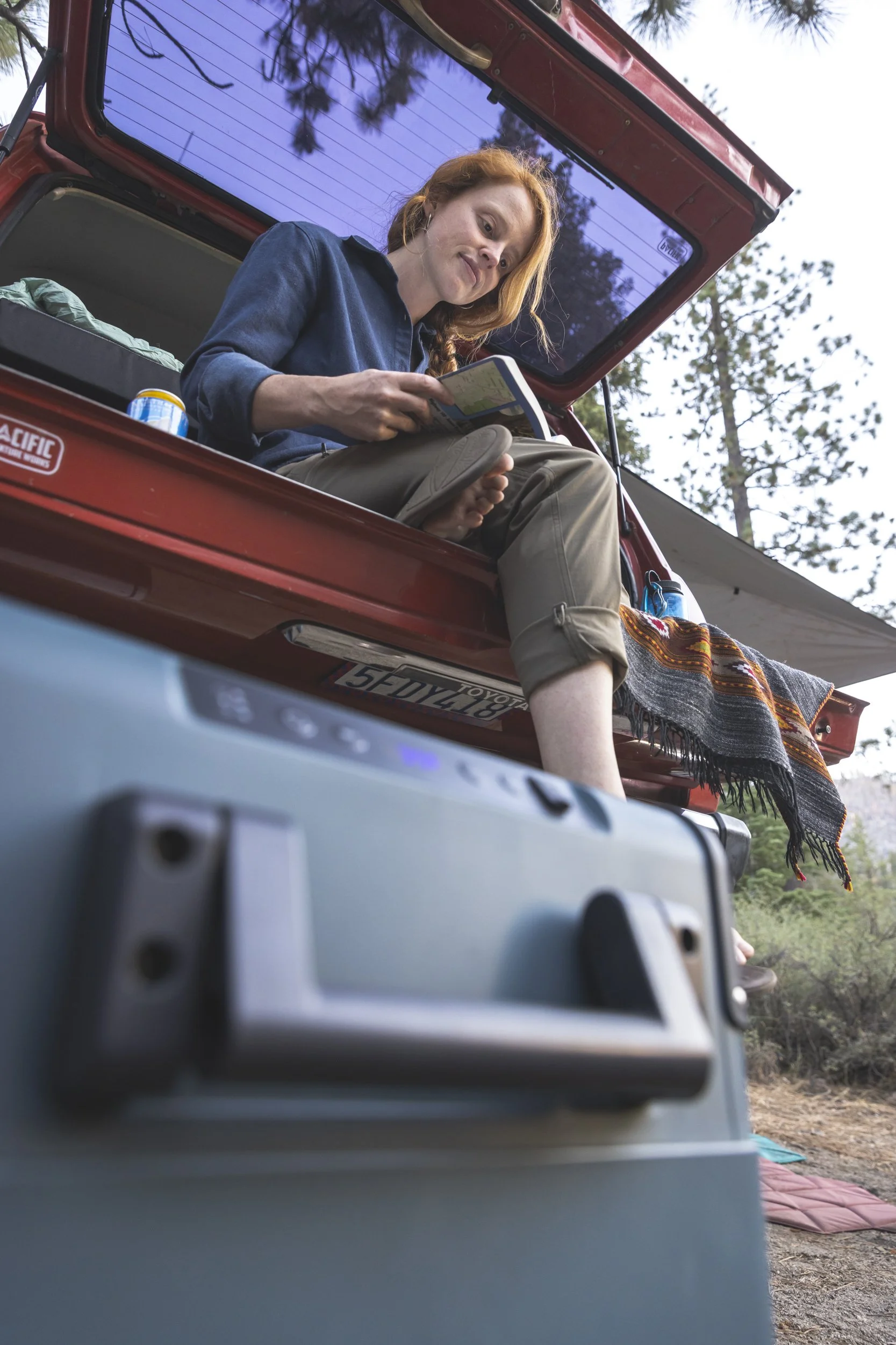 Woman sitting on the edge of a red vintage camper trunk, reading a guidebook with a blanket beside her and a Dometic cooler on the ground in the foreground, outdoors with trees in the background, photographed by Fieldborn Creative.