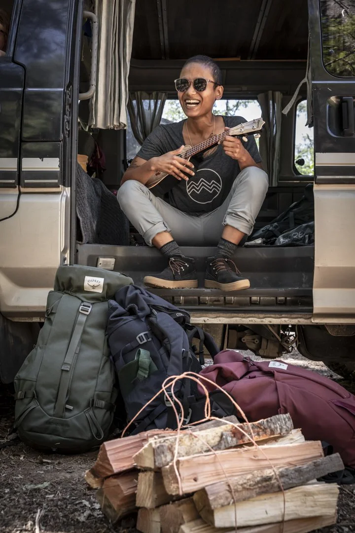 Person sitting in open side door of camper van holding ukulele, smiling, with backpacks and camping gear, photographed by Fieldborn Creative