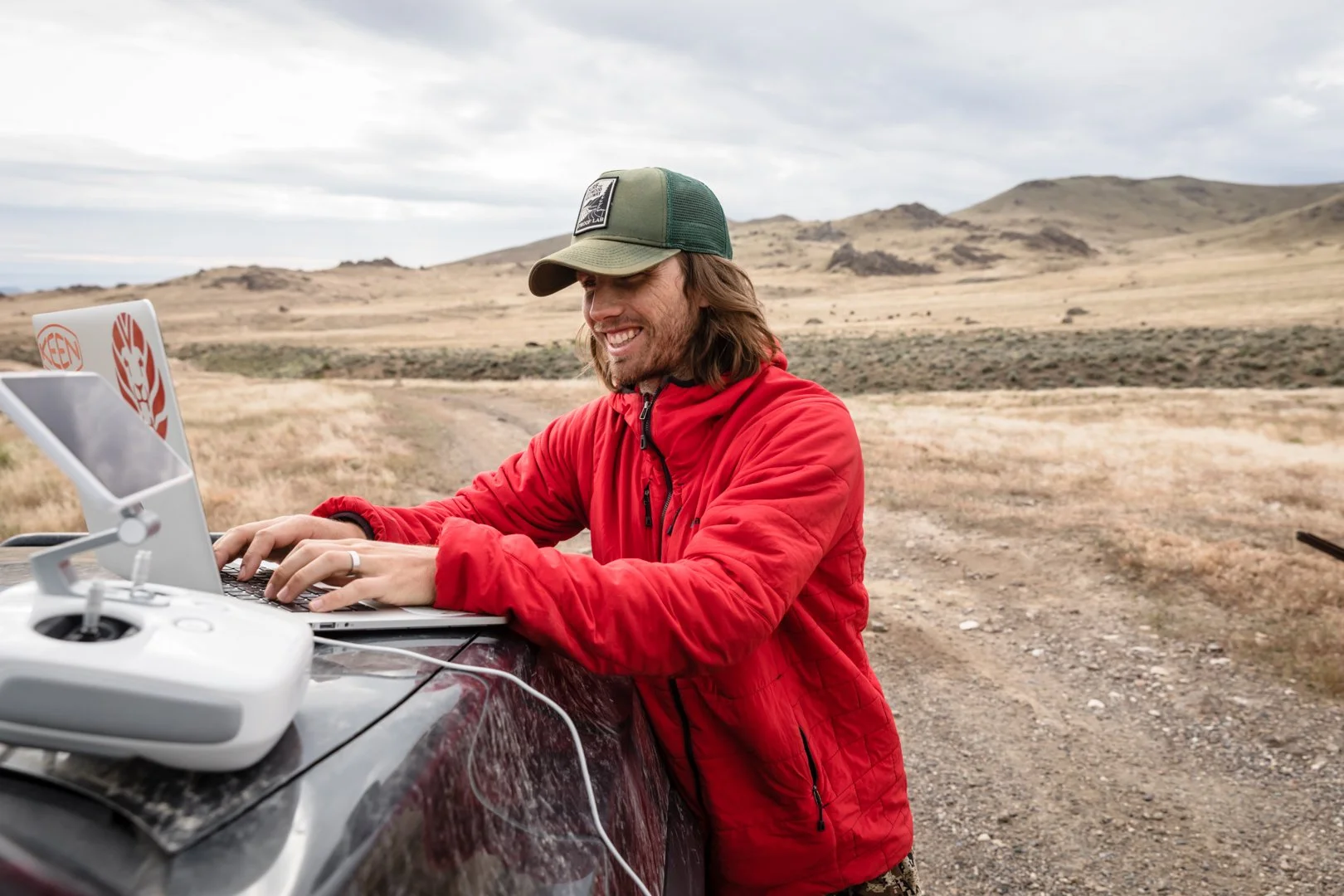 Behind-the-scenes photo of a man using a laptop on the hood of a truck with a drone controller in the foreground, photographed by Fieldborn Creative in the field.