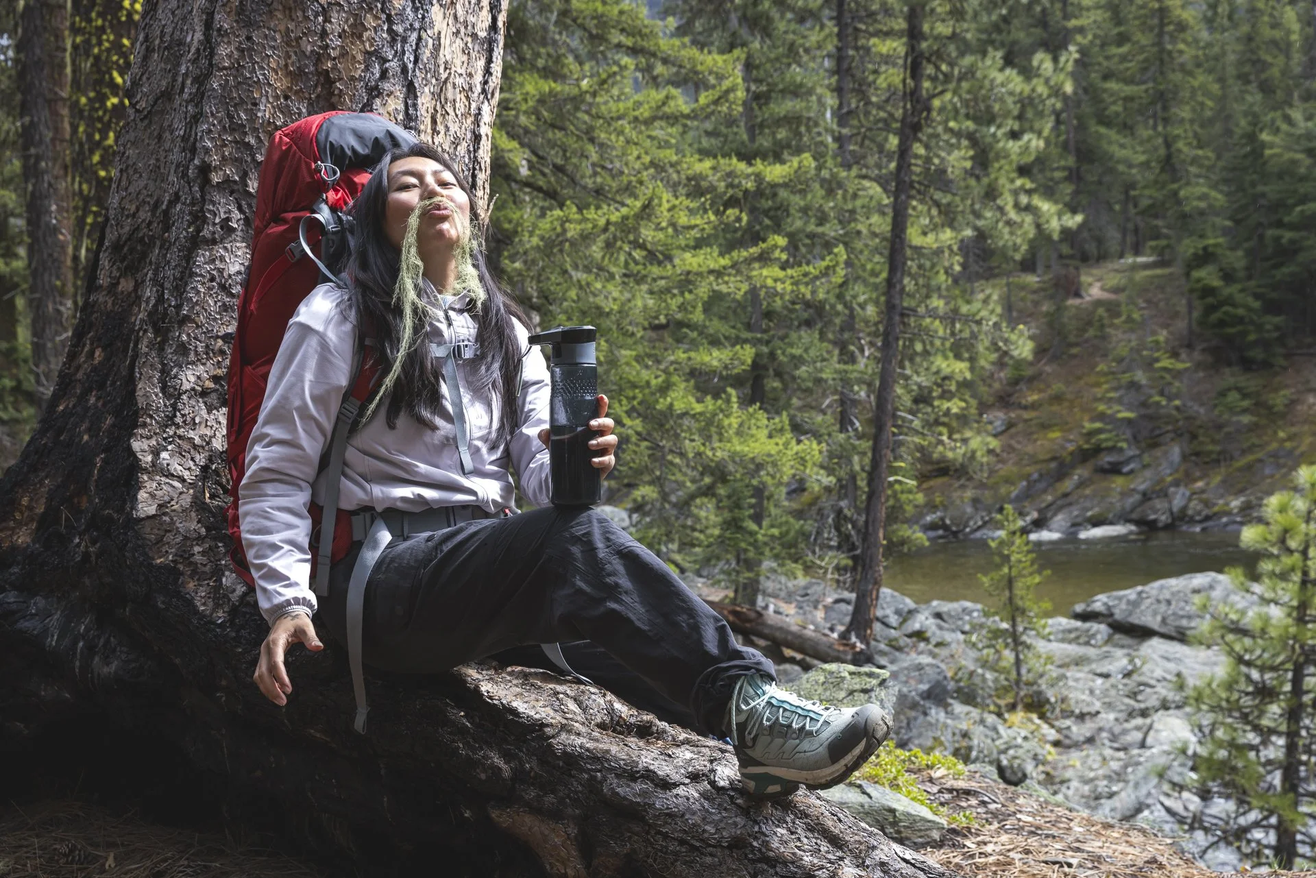 A woman with a backpack sitting against a tree in a forest, holding a water bottle and smiling.