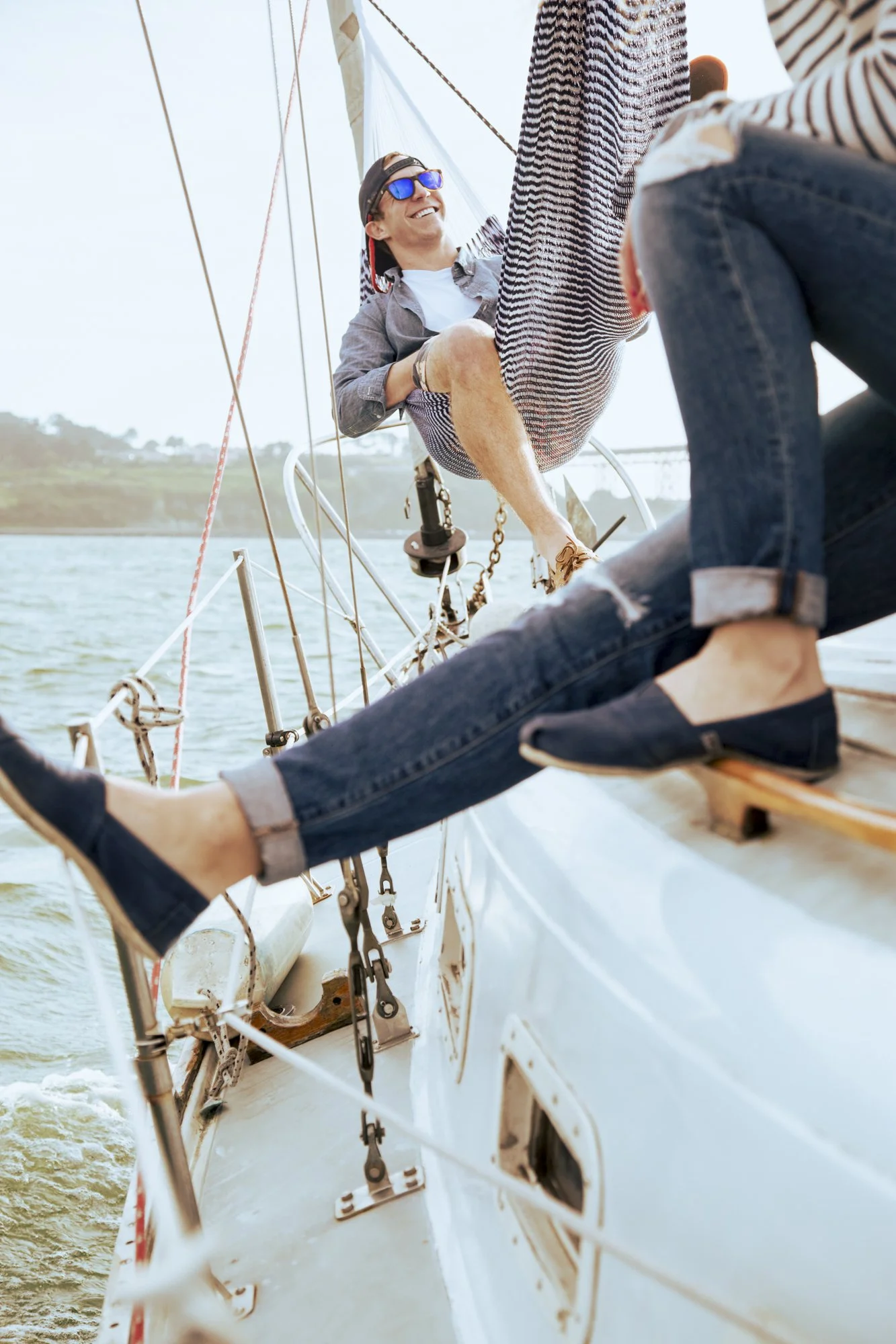 A man with sunglasses and a backward cap relaxing on a sailboat with two other people, smiling and enjoying the outdoors, with water and distant land visible. Commercial lifestyle production by Fieldborn Creative.
