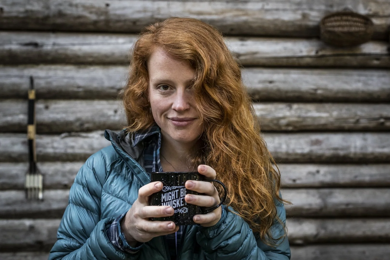 Woman with red hair wearing blue jacket holding black mug reading “Might be whiskey” in front of wooden log wall, photographed by Fieldborn Creative