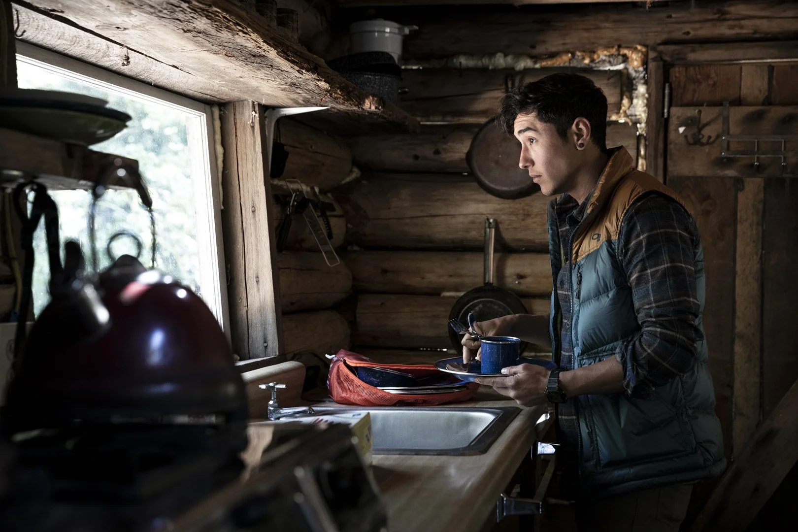 Young man in plaid shirt and puffy vest holding plate with blue mug in rustic cabin kitchen, photographed by Fieldborn Creative