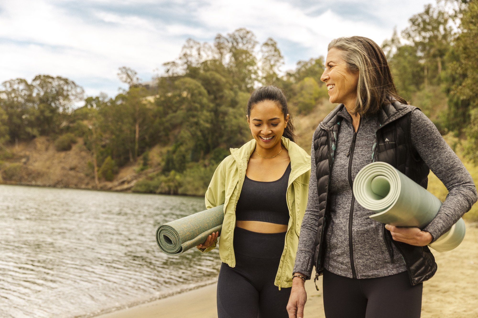 Two women walking along the shore carrying yoga mats, with trees and a cloudy sky in the background. Commercial lifestyle production by Fieldborn Creative.