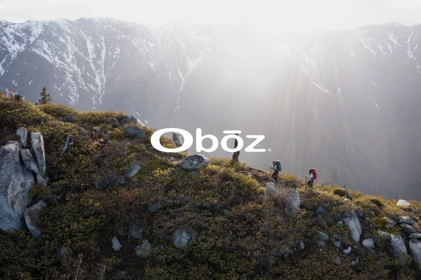 Hikers walking on a mountain trail with backpacks, overlooking a valley with snow-capped mountains in the distance and sunlight shining through the sky.