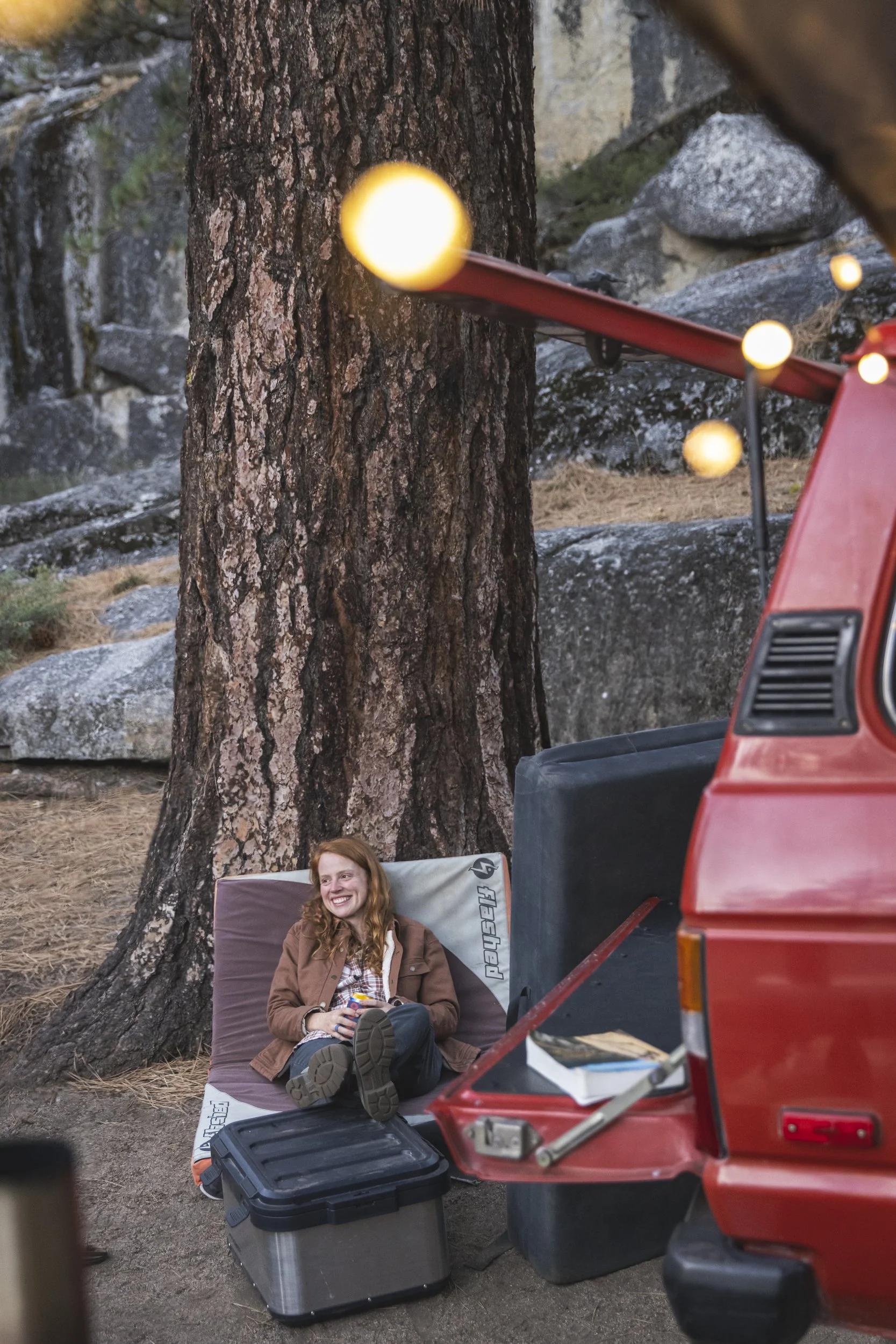 Red-headed woman sitting on a bouldering crash pad, drinking a beer with her feet on a Dometic storage case near a tailgate in a California forest, photographed by Fieldborn Creative.