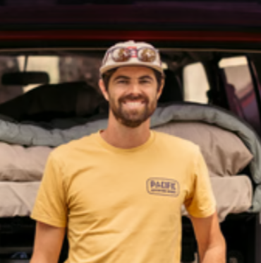 Man with a beard smiling, wearing a yellow T-shirt and a cap with goggles, standing in front of an open vehicle trunk with camping gear.