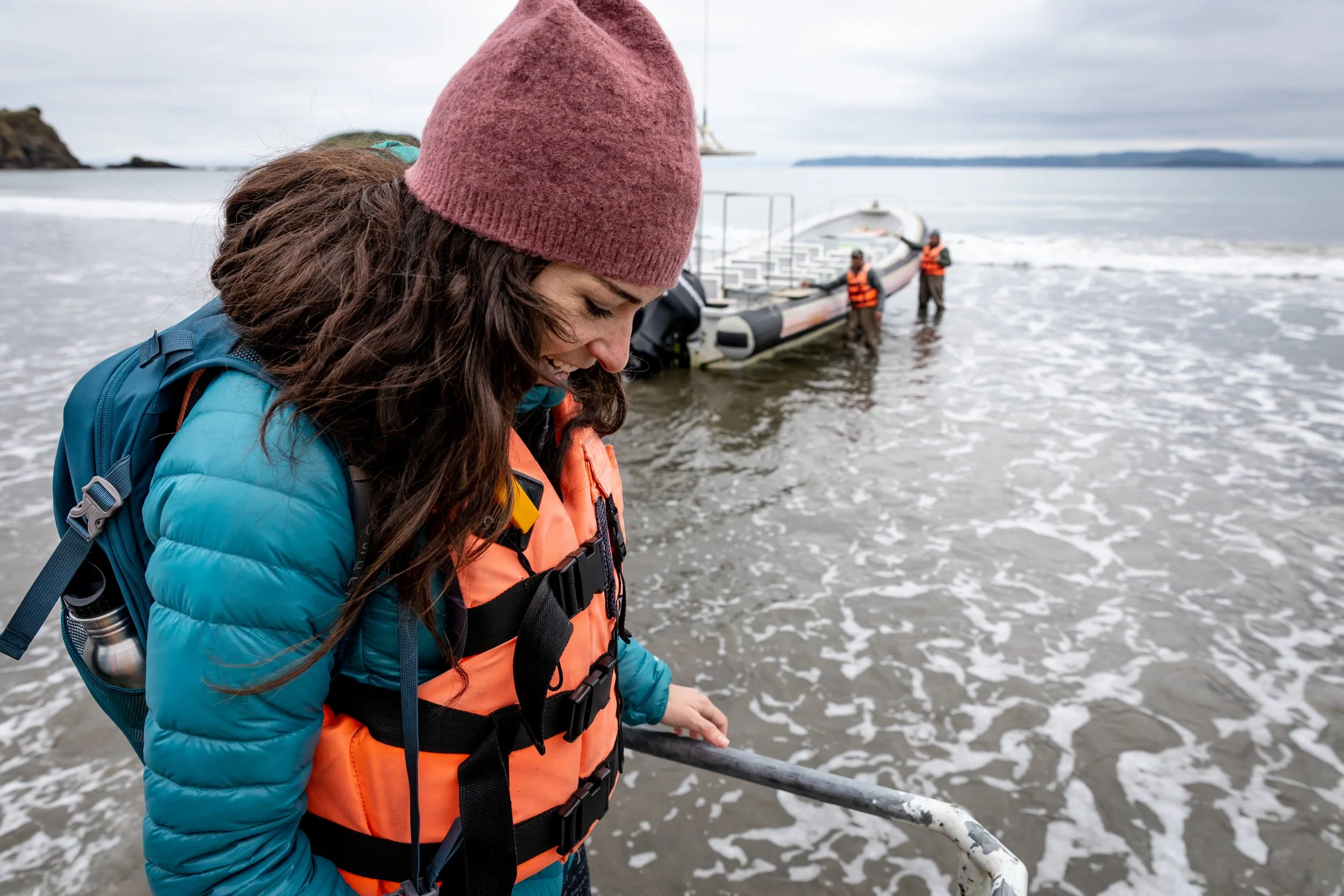 A woman with long brown hair wearing a pink knit beanie, blue jacket, and orange life vest stands on the beach holding a metal railing, with a boat and two people in life vests in the water behind her. Commercial production by Fieldborn Creative.
