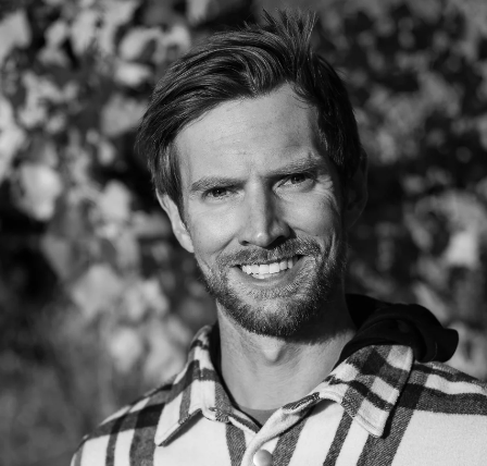 A smiling man with short hair and a beard wearing a plaid shirt outdoors.