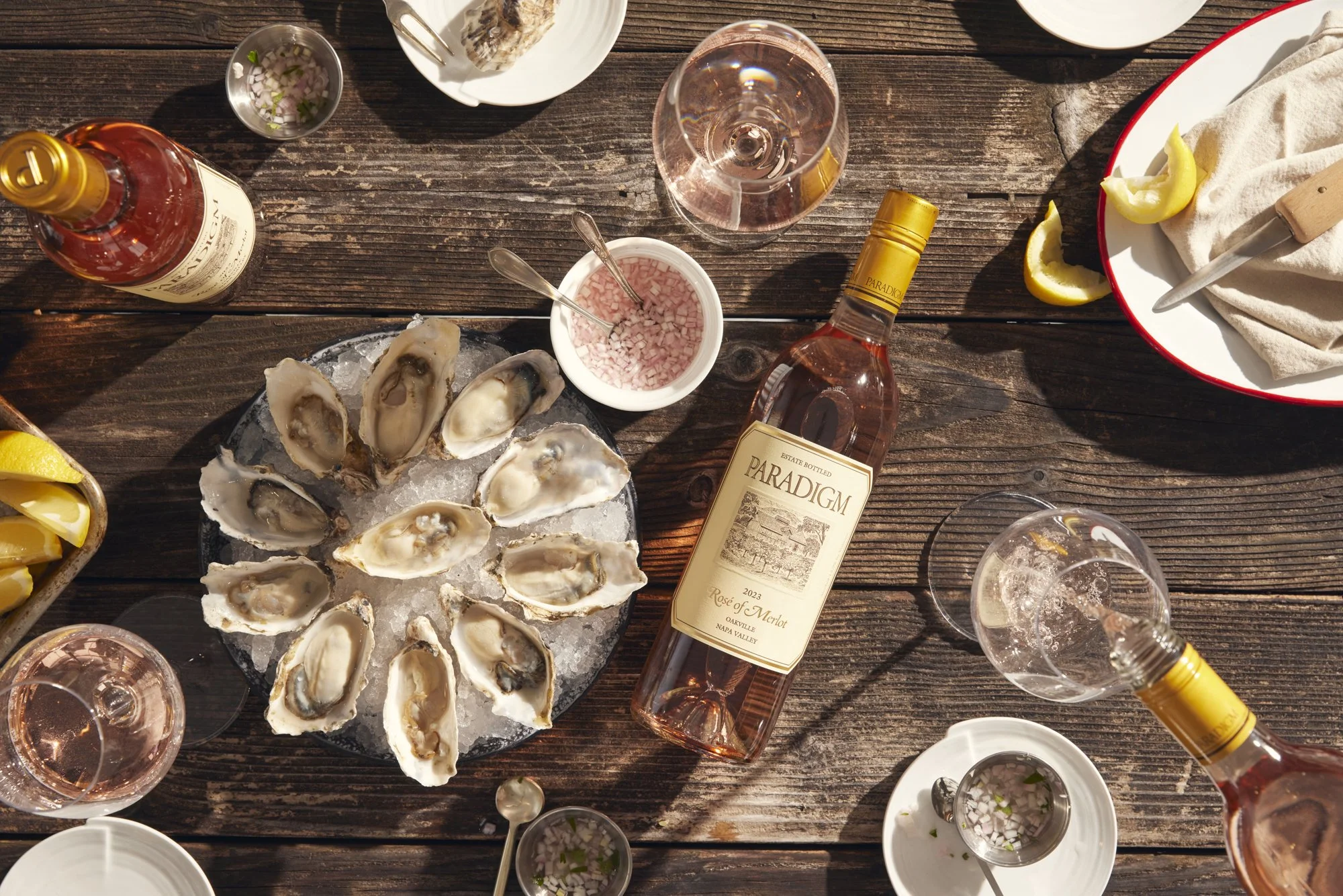 A top-down view of a seafood dining table with oysters on a bed of ice, two bottles of rosé wine, glasses of pink wine, small bowls of chopped onions and condiments, lemon wedges, and various white dishes on a wooden surface. 