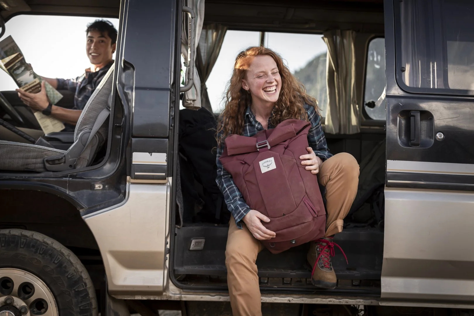 Woman with red hair holding maroon backpack sitting in open camper van door, man in driver’s seat holding map, photographed by Fieldborn Creative