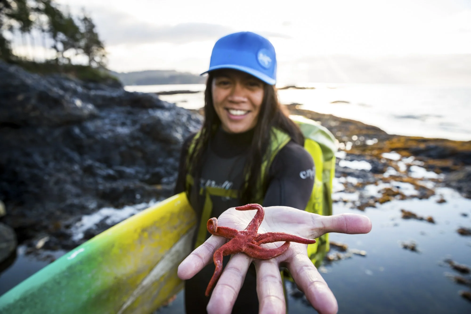 A smiling woman in outdoor gear holding a red starfish over a rocky shoreline with water and trees in the background. Commercial lifestyle production by Fieldborn Creative.
