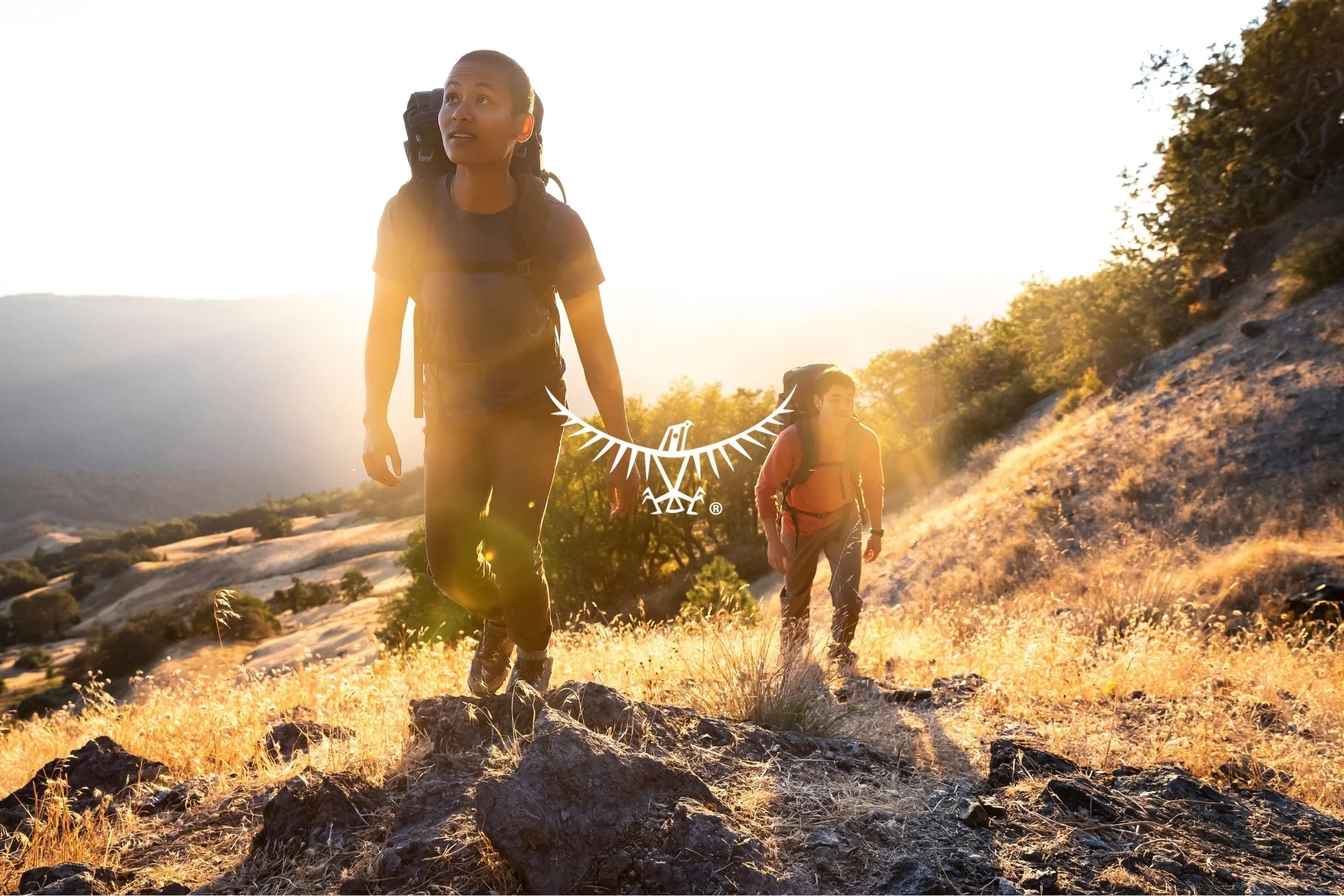 Two hikers walking on rocky trail in a mountainous area during sunset, with trees and hills in background.