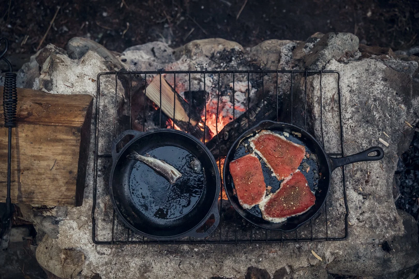 Cast iron skillet with fresh caught fish cooking over campfire in forest, Osprey Archeon pack nearby, shot by Fieldborn Creative
