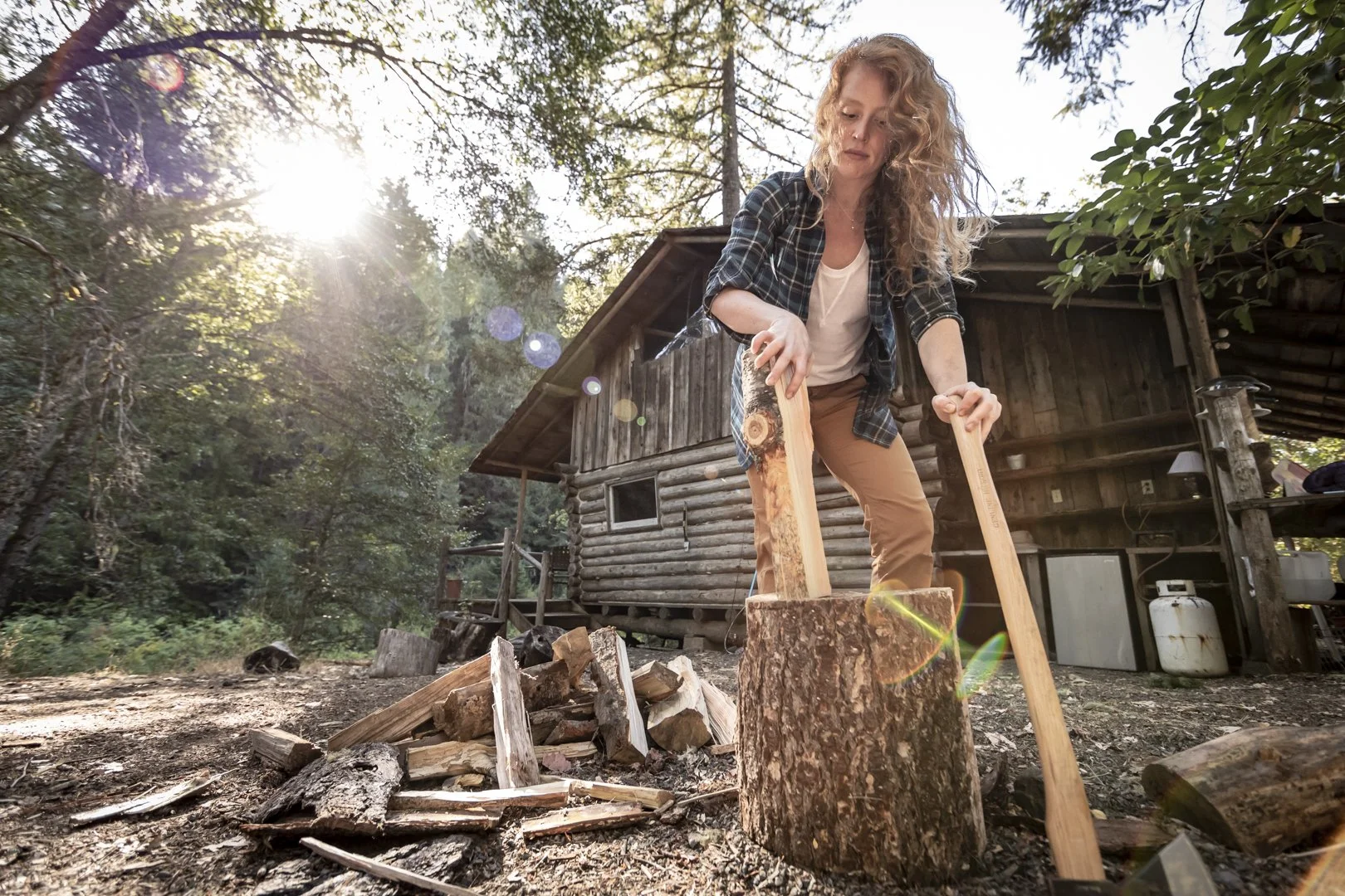 Woman chopping wood with axe outside rustic cabin in forest, sunlight filtering through trees, photographed by Fieldborn Creative