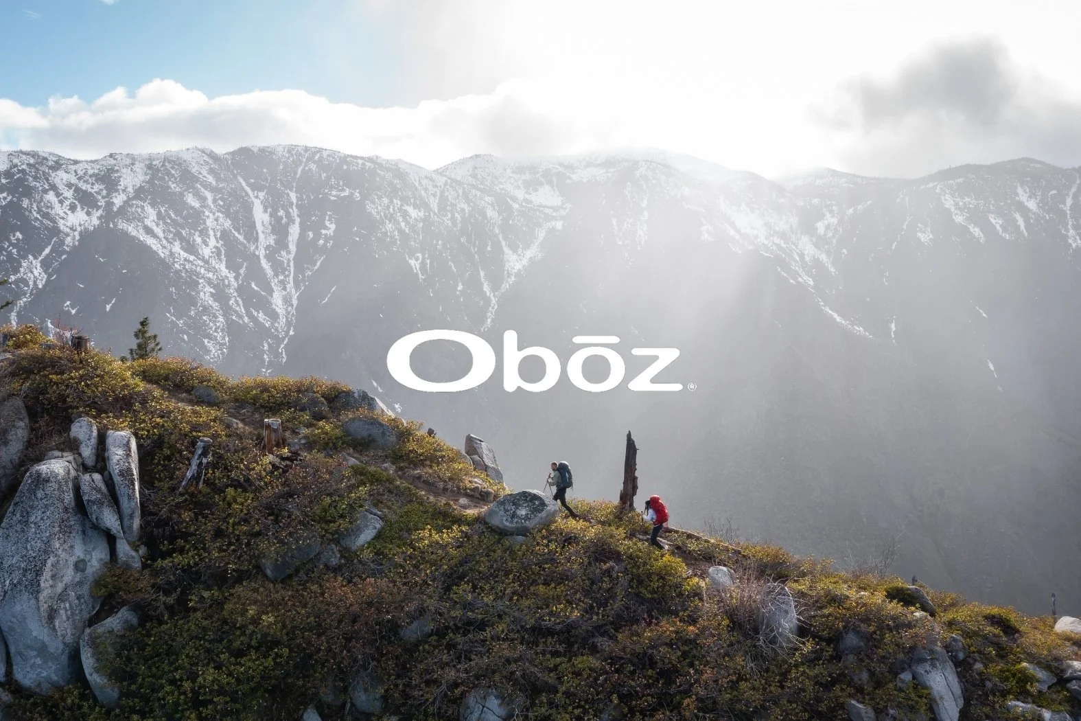 Two hikers with backpacks walking along a mountain trail with a backdrop of snow-capped mountains and cloudy sky. Commercial lifestyle production by Fieldborn Creative.