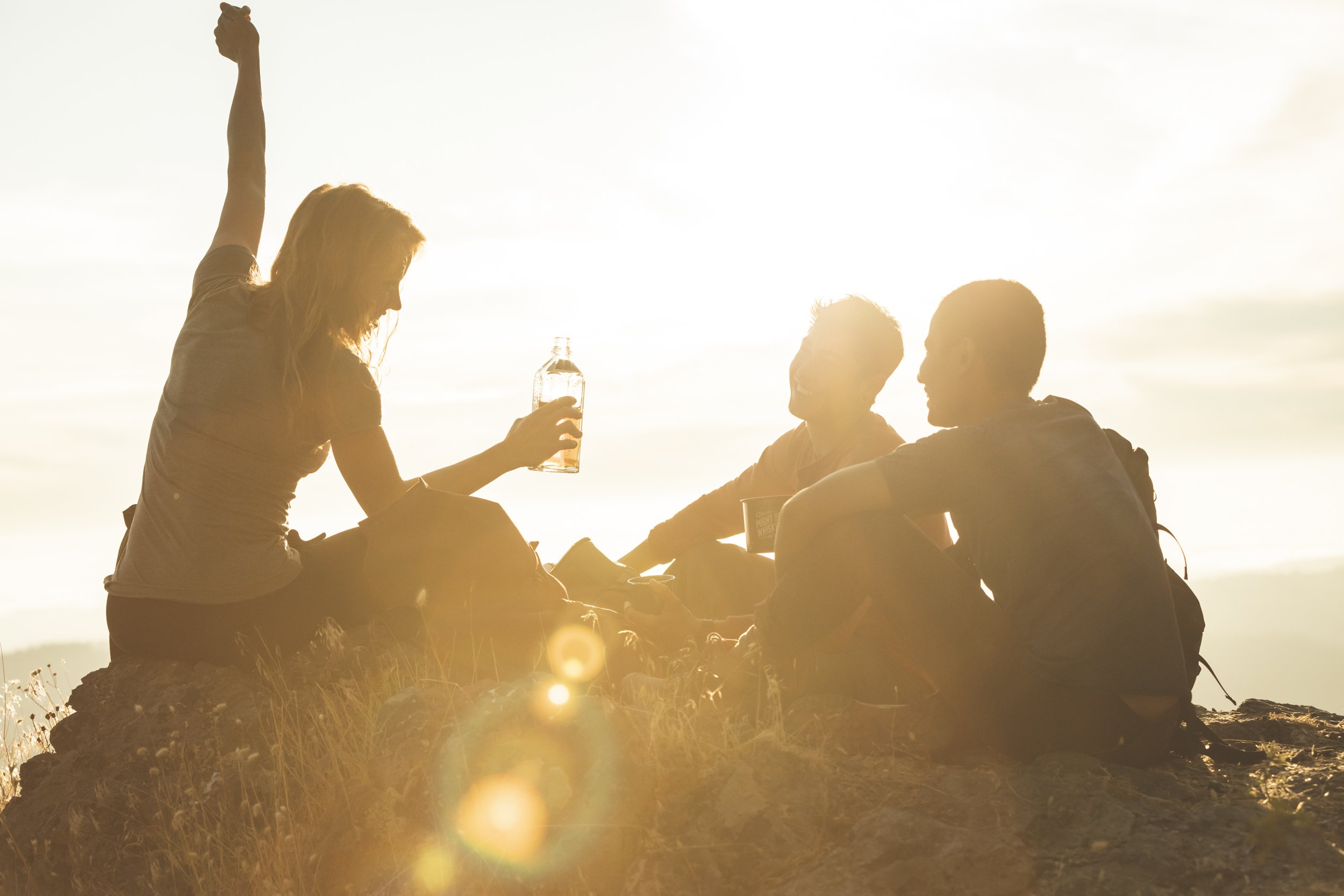 Three friends—one man and two woman—sitting on rock outdoors enjoying drinks and conversation at sunset, photographed by Fieldborn Creative