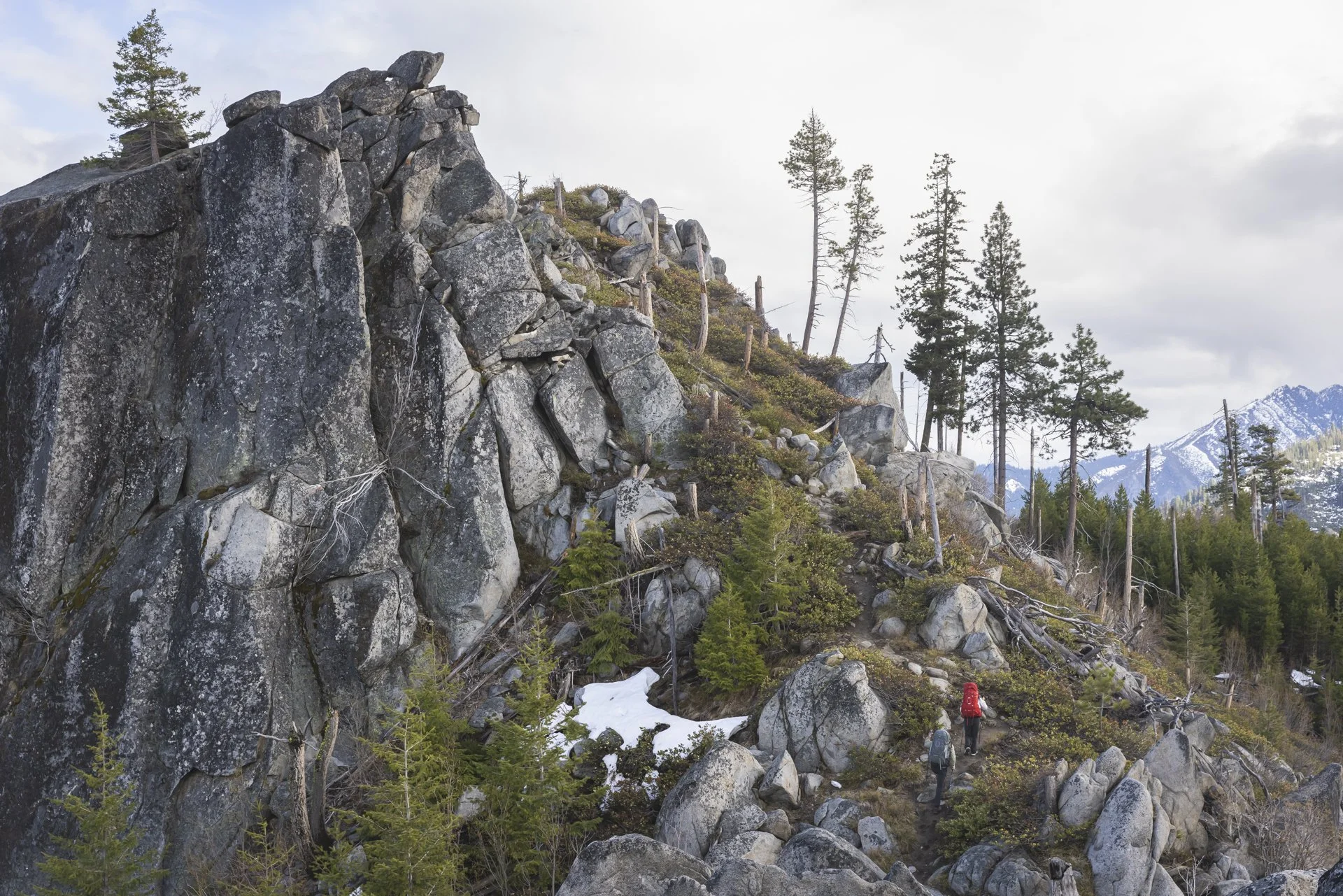 Two hikers walking along a rocky mountain trail surrounded by trees and snow patches, with distant snow-capped mountains in the background.