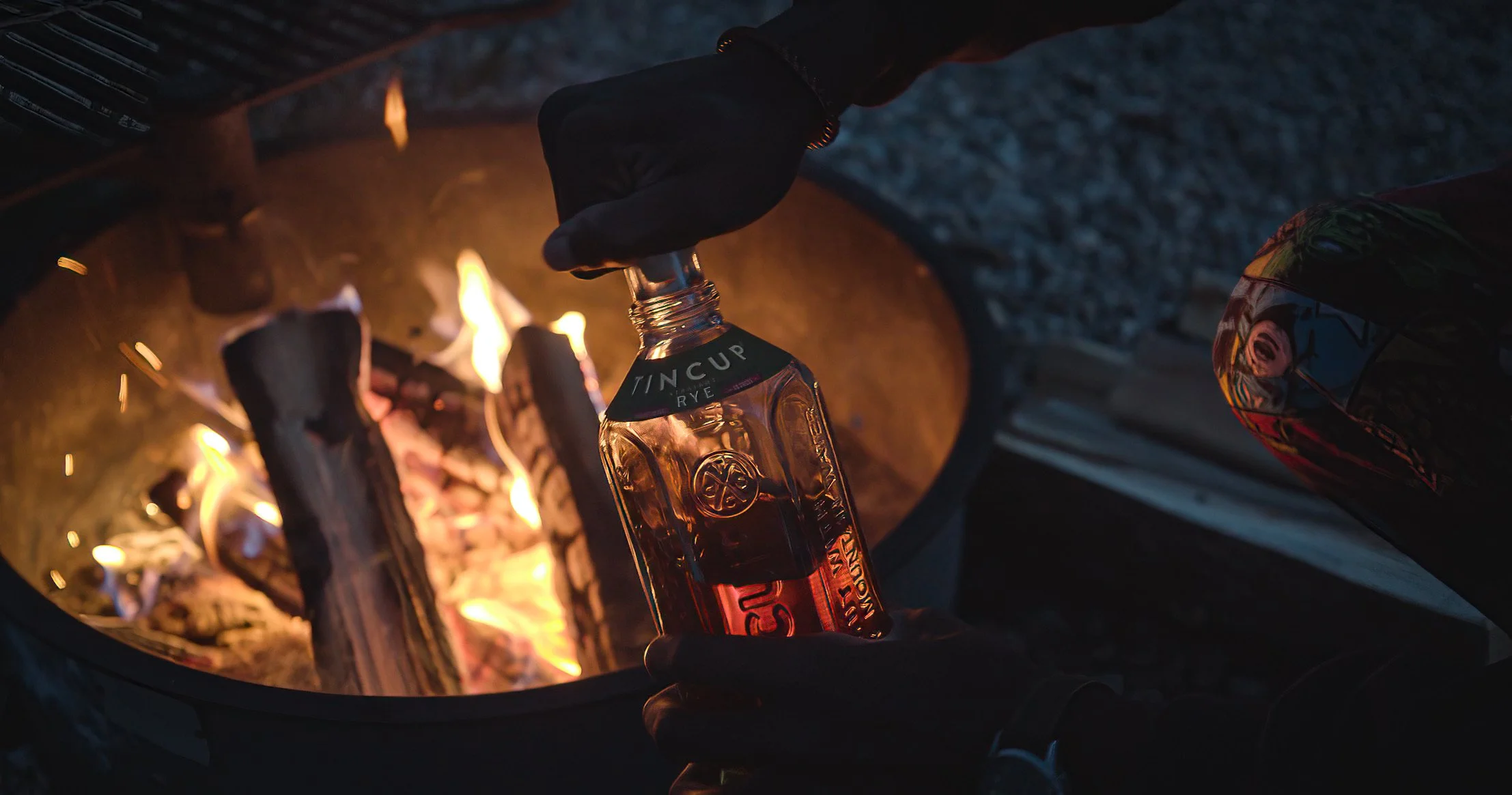 Person wearing black gloves is holding a bottle of Tincup whiskey over a campfire with burning logs.