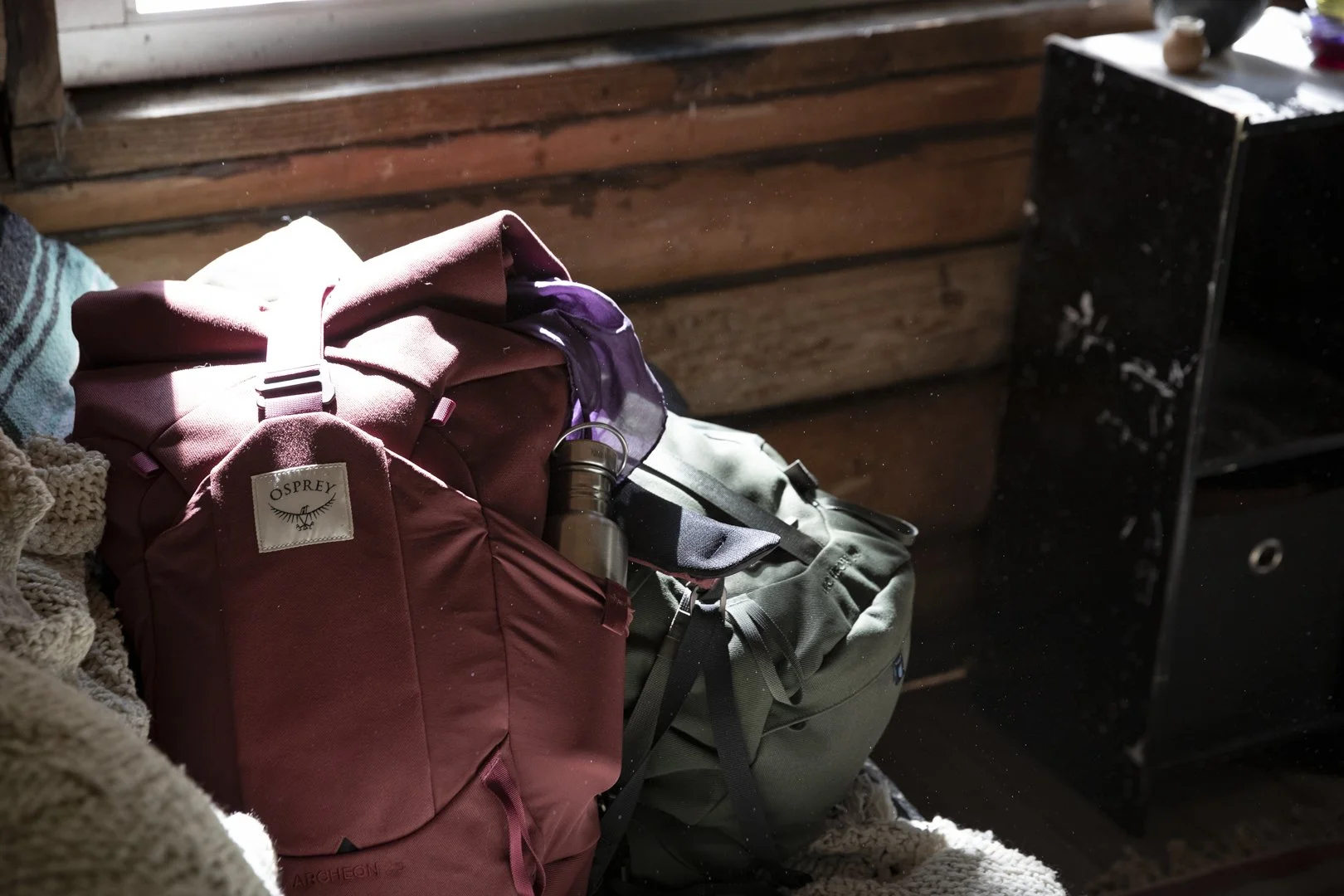 Red Osprey Archeon backpack with water bottle on blanket inside wooden cabin, photographed by Fieldborn Creative