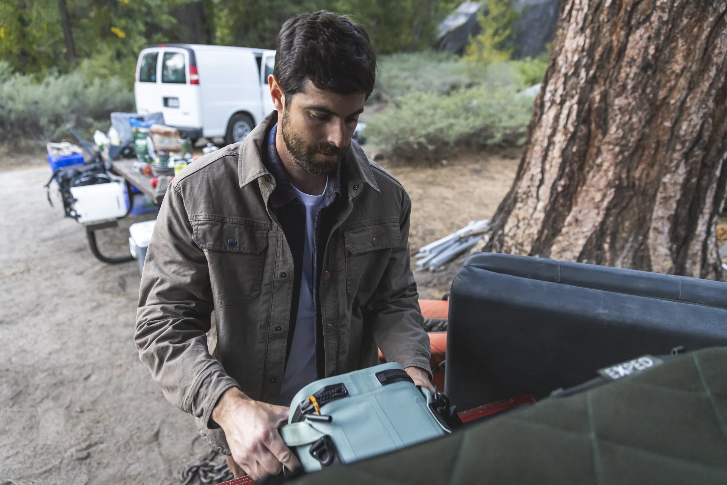 Man organizing camping gear outdoors near a large tree, placing a Dometic cooler in a truck at a campground, with a white van and picnic table in the background, photographed by Fieldborn Creative.
