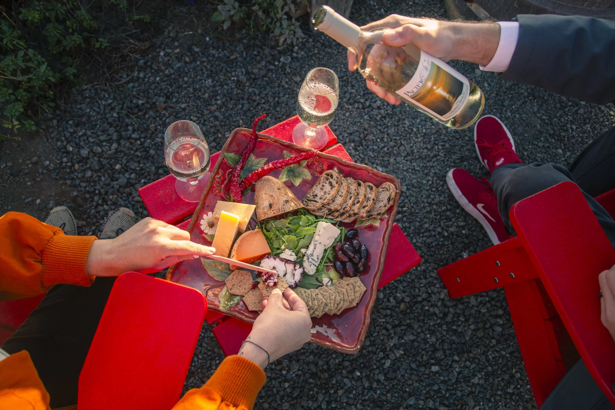 People having a picnic outdoors on a red table with cheese, crackers, bread, grapes, and chilies, while pouring wine into glasses. Commercial lifestyle production by Fieldborn Creative.