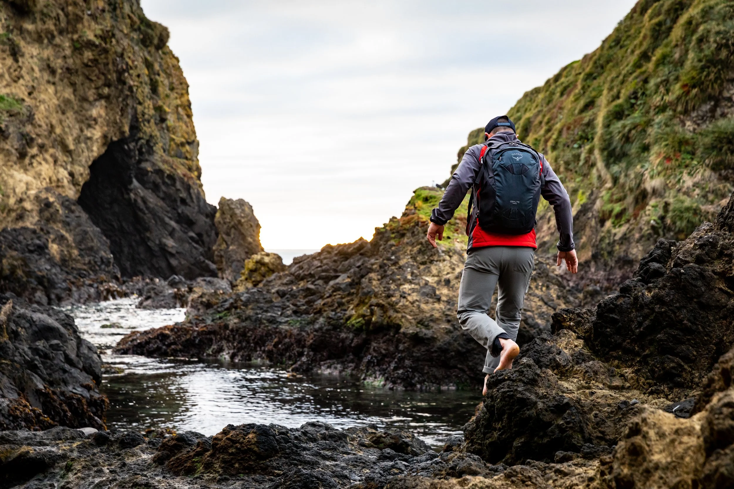 A person with a black cap, gray jacket with red accents, and gray pants walking barefoot through rocky terrain beside a small stream, flanked by moss-covered cliffs on both sides. Commercial lifestyle production by Fieldborn Creative.
