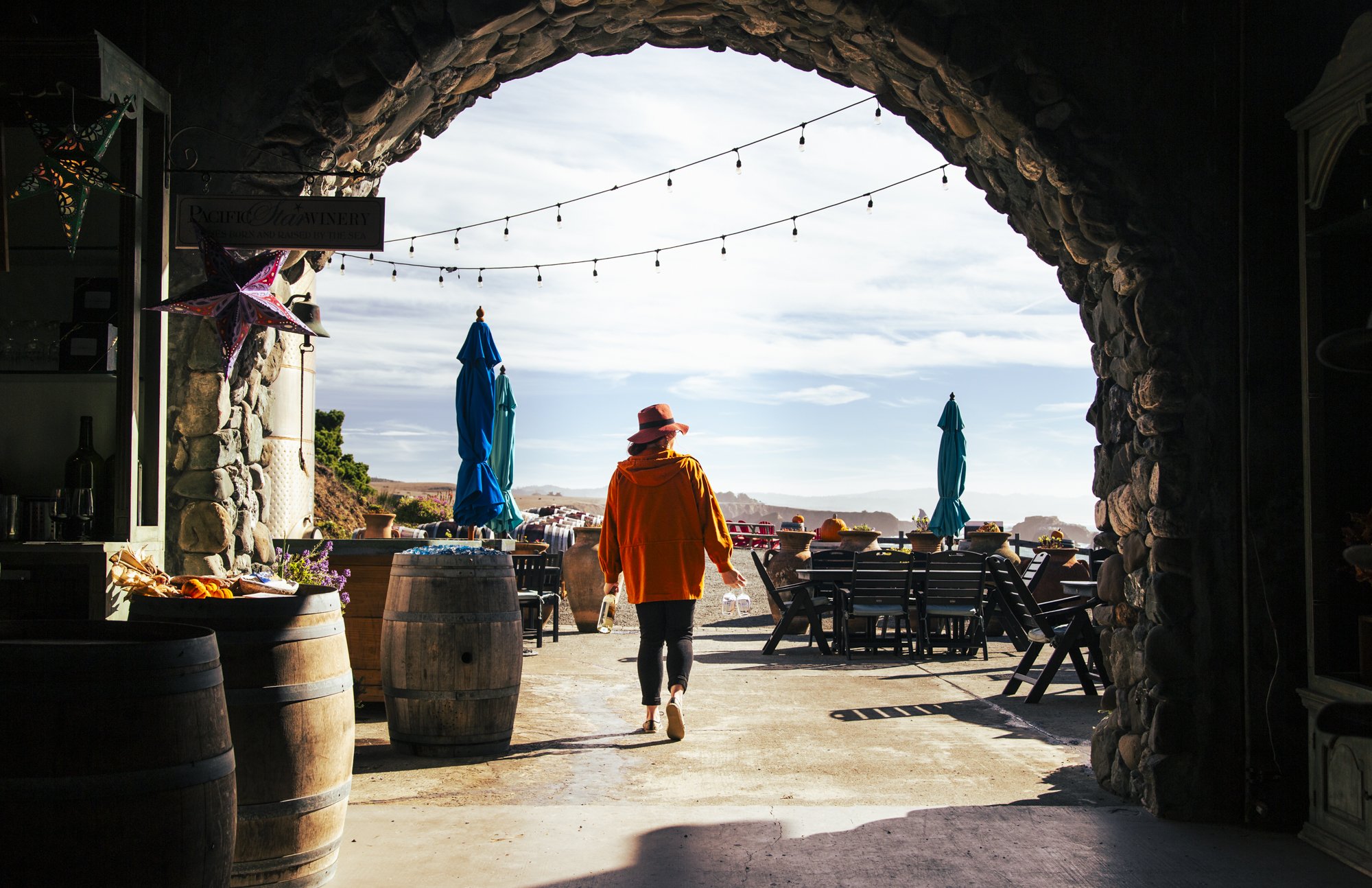 A person with a pink hat, orange jacket, and black pants walking away with a water bottle in each hand through an outdoor patio area under a stone archway, with string lights overhead, blue umbrellas, chairs, tables, wine barrels, and decorative item