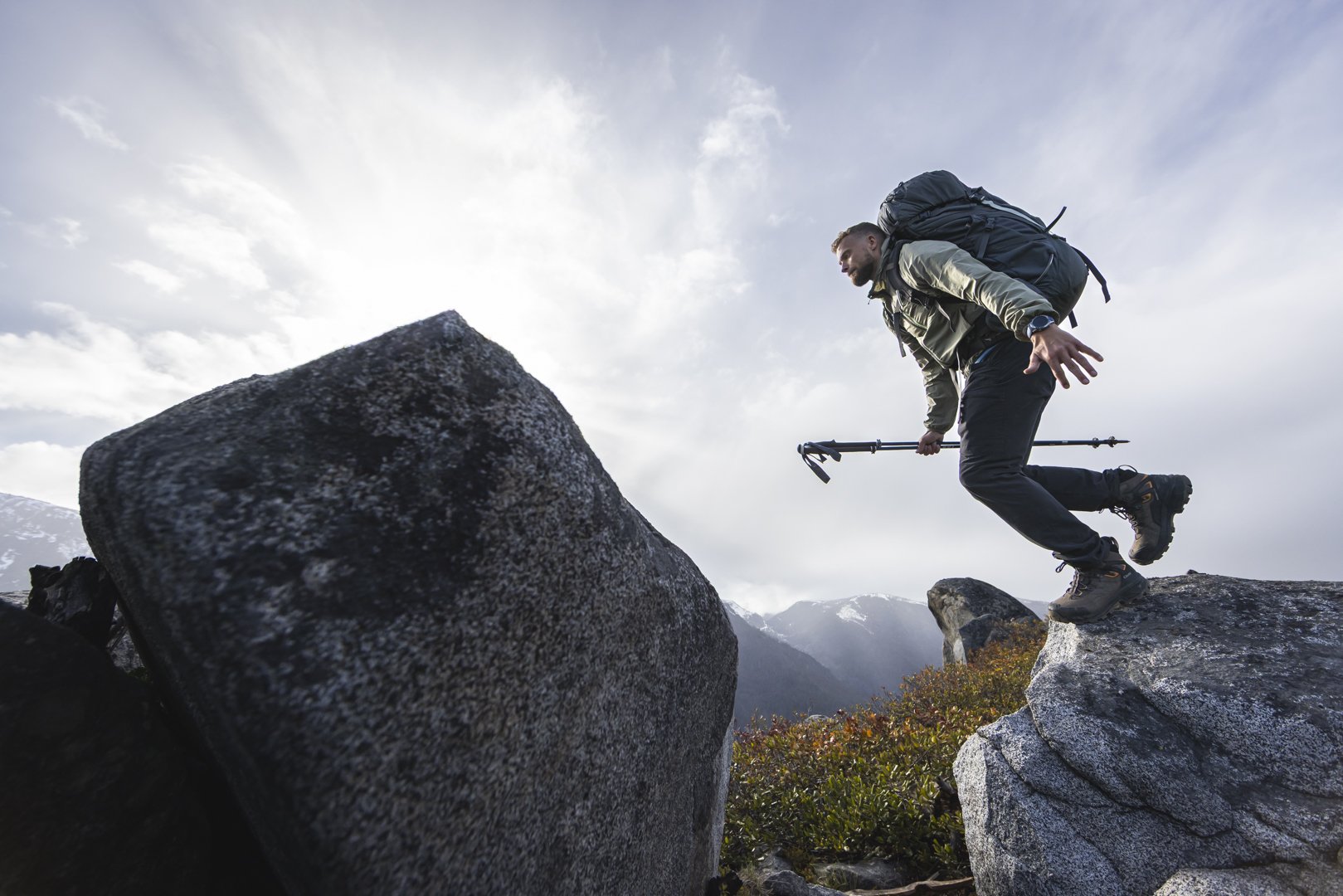 A man with a backpack and trekking pole balancing on a rock in a mountainous landscape.
