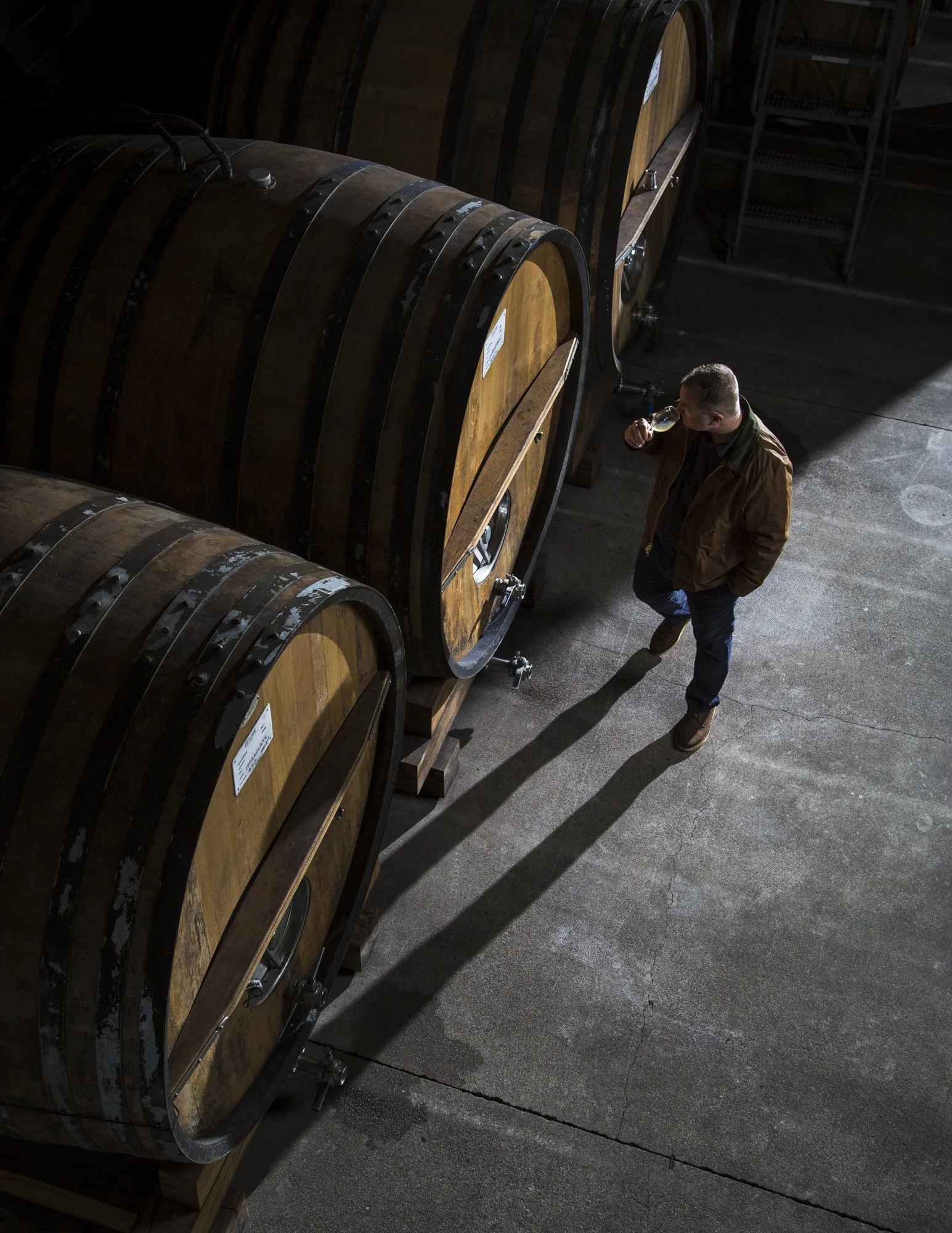 A man tasting wine in a winery surrounded by large wooden wine barrels. Commercial lifestyle production by Fieldborn Creative.