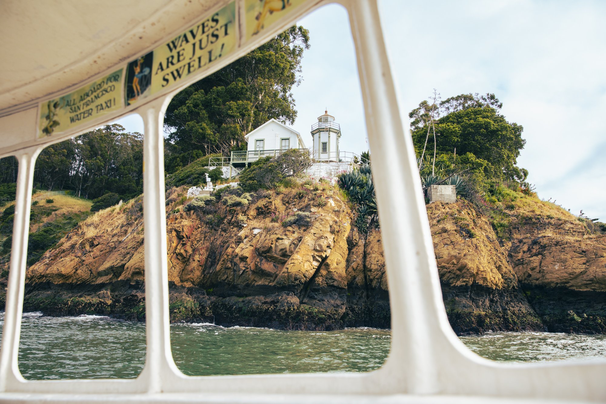 View of a lighthouse and white building on rocky hillside surrounded by green trees, seen through the railings of a boat, with water at the bottom of the image. Commercial lifestyle production by Fieldborn Creative.