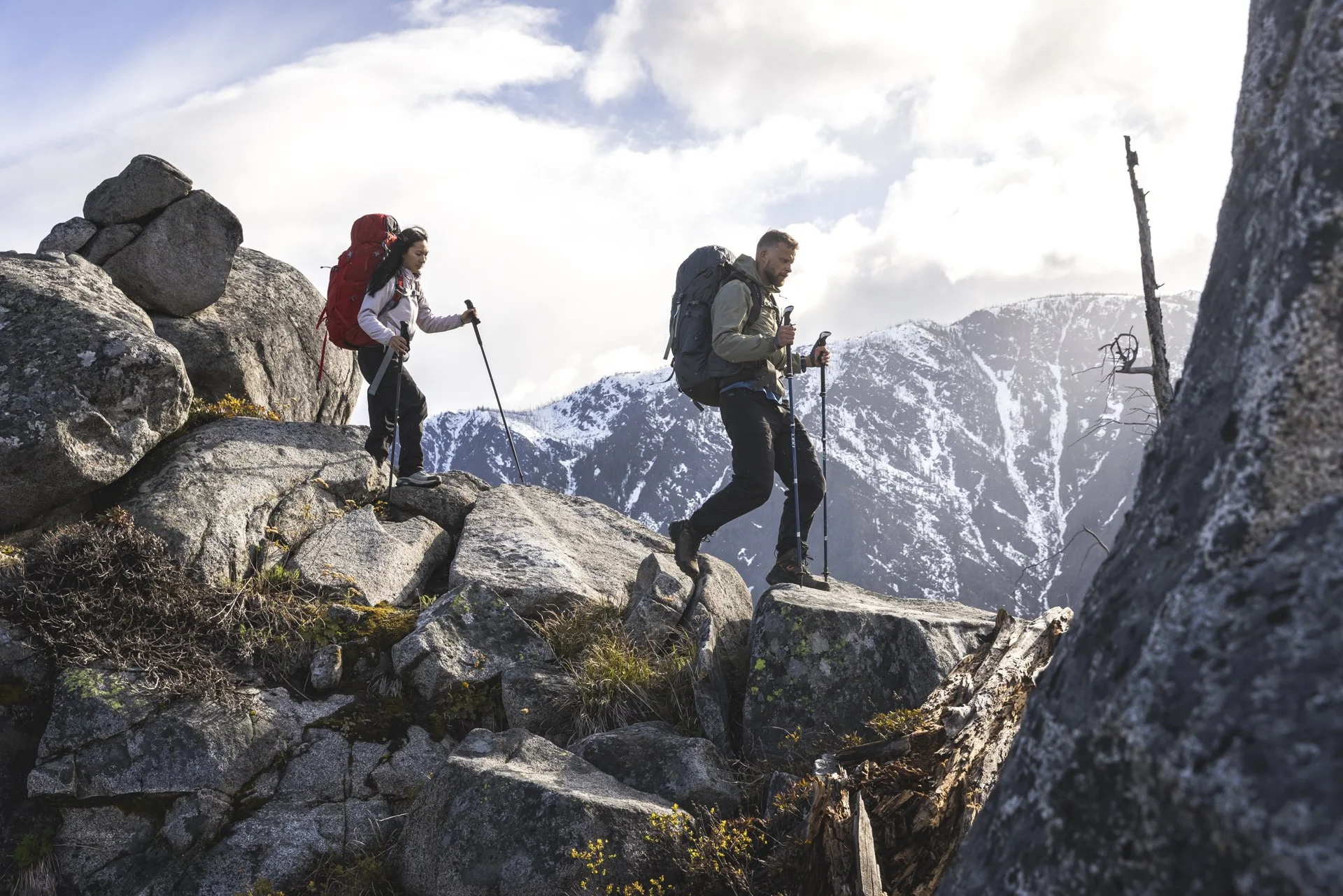 Two hikers in outdoor gear hiking over rocky terrain with snow-capped mountains in the background.