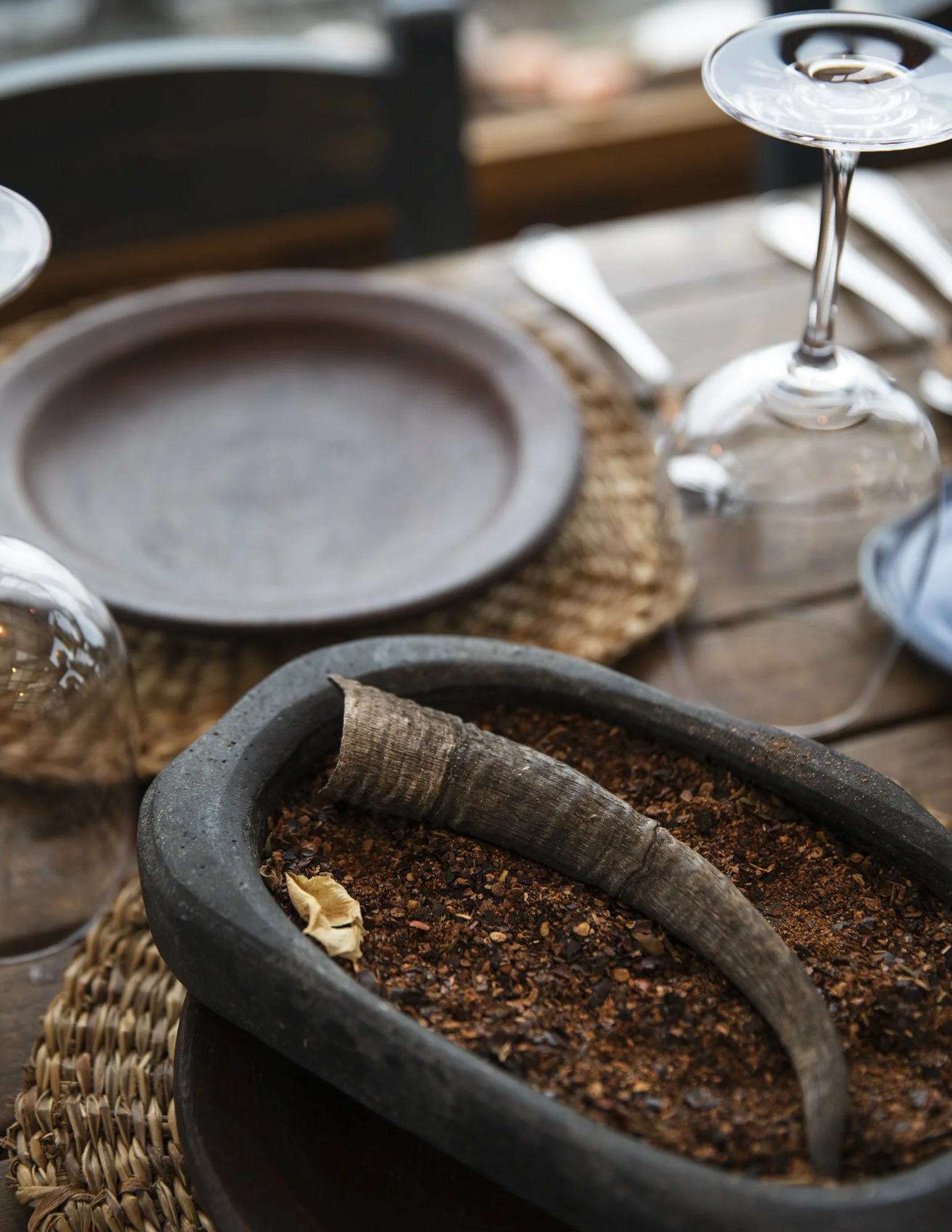 A black flower pot filled with soil and a used dried brown banana leaf. There is a conical object, possibly a dried seed or pod, in the soil. The pot is placed on a woven surface, and the background shows a wooden surface with various kitchen utensil