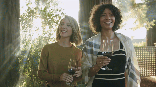 Two women smiling and holding glasses of red wine outdoors, with sunlight filtering through trees in the background. Commercial lifestyle production by Fieldborn Creative.