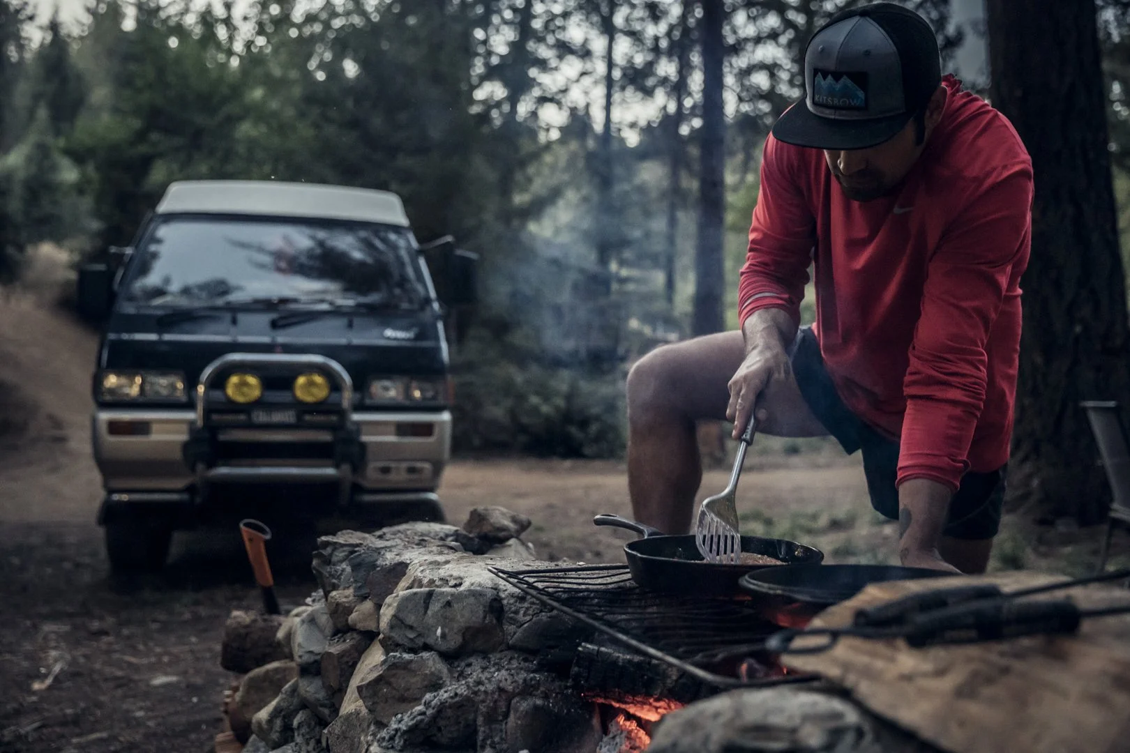 Man in red sun shirt cooking fish on campfire in forest with camper van, photographed by Fieldborn Creative for Osprey Packs