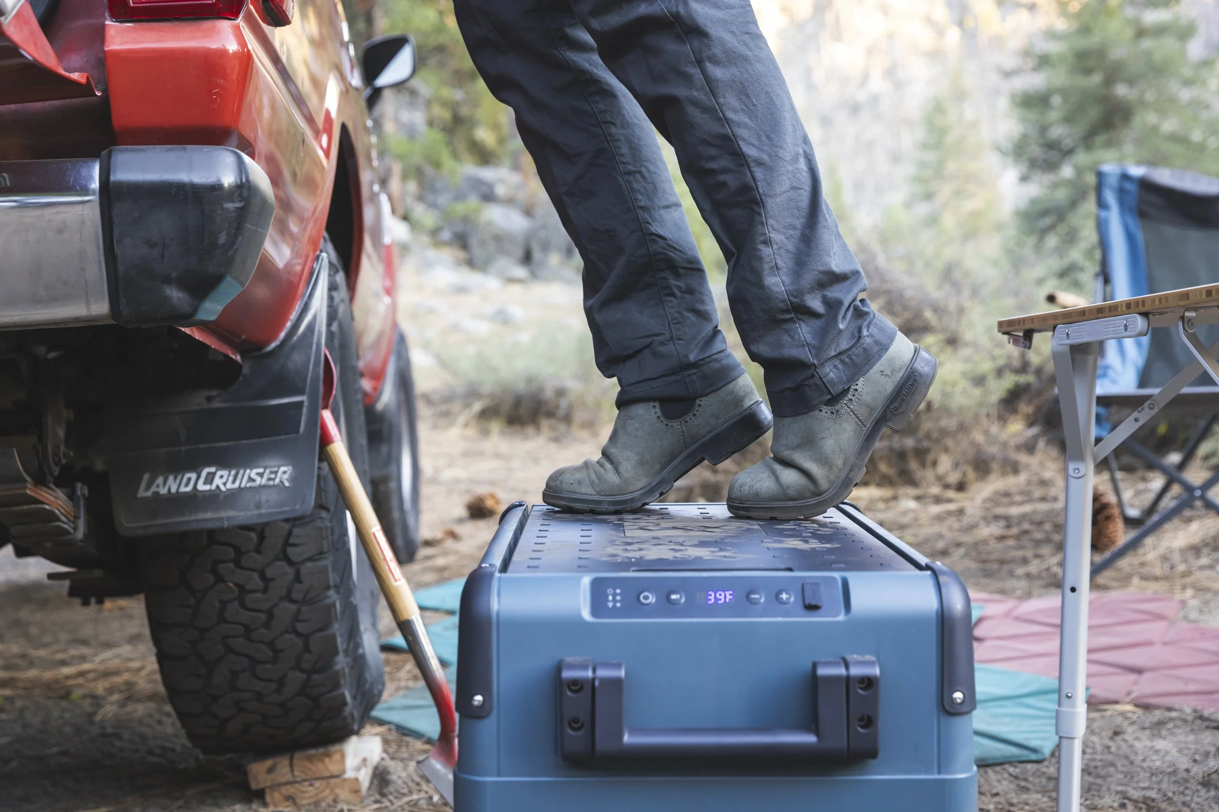 Person standing on a portable Dometic electric cooler at a campsite near a red Toyota Land Cruiser, wearing black pants and green boots, with camping chairs and gear visible in the background, photographed by Fieldborn Creative.