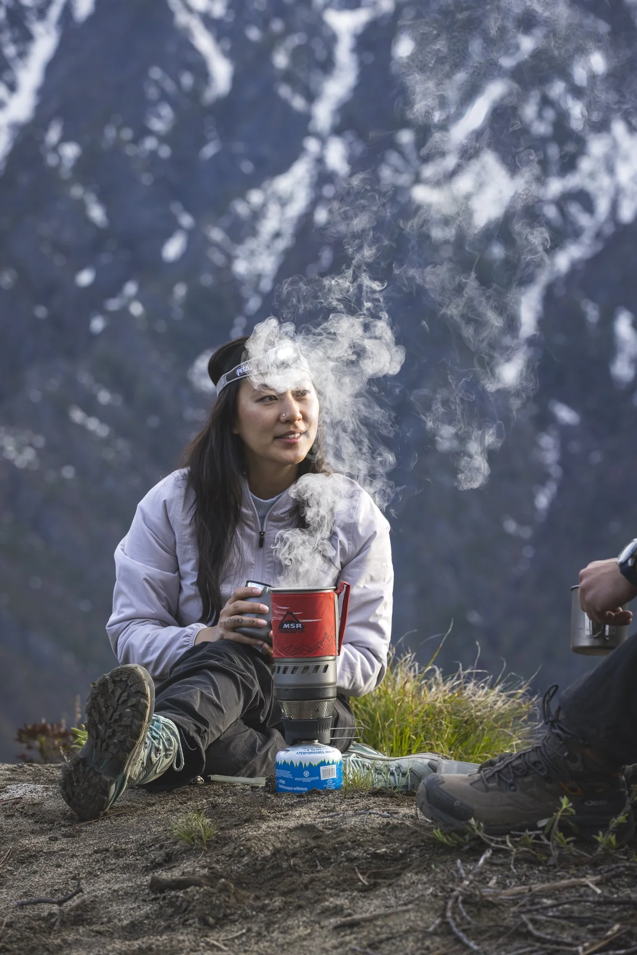 A woman in outdoor gear sitting on a rock, brewing hot beverages with a portable stove, with mountains and trees in the background.