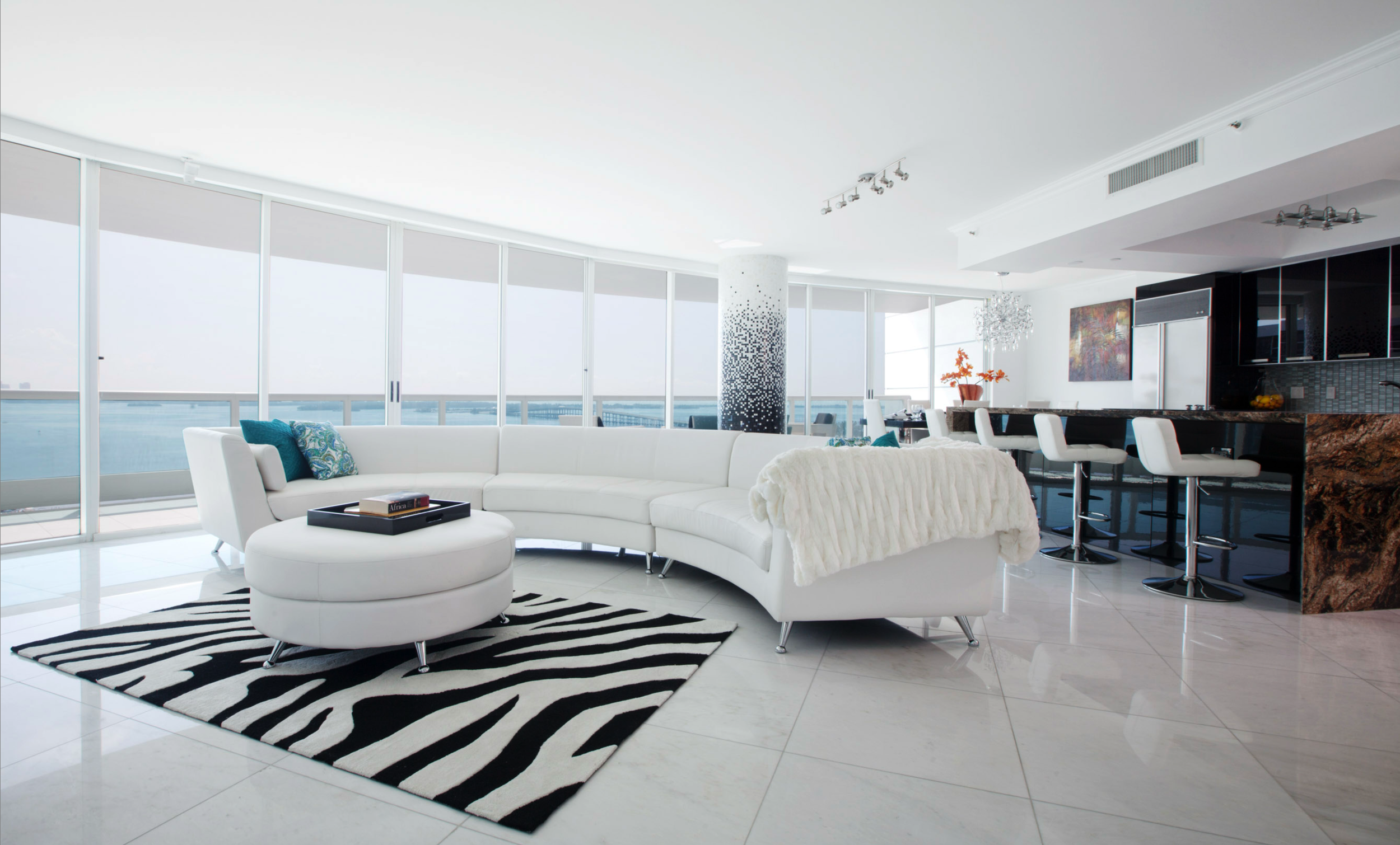 Modern living room with white curved sofa, black and white zebra rug, large windows, and kitchen in the background. Commercial lifestyle production by Fieldborn Creative.