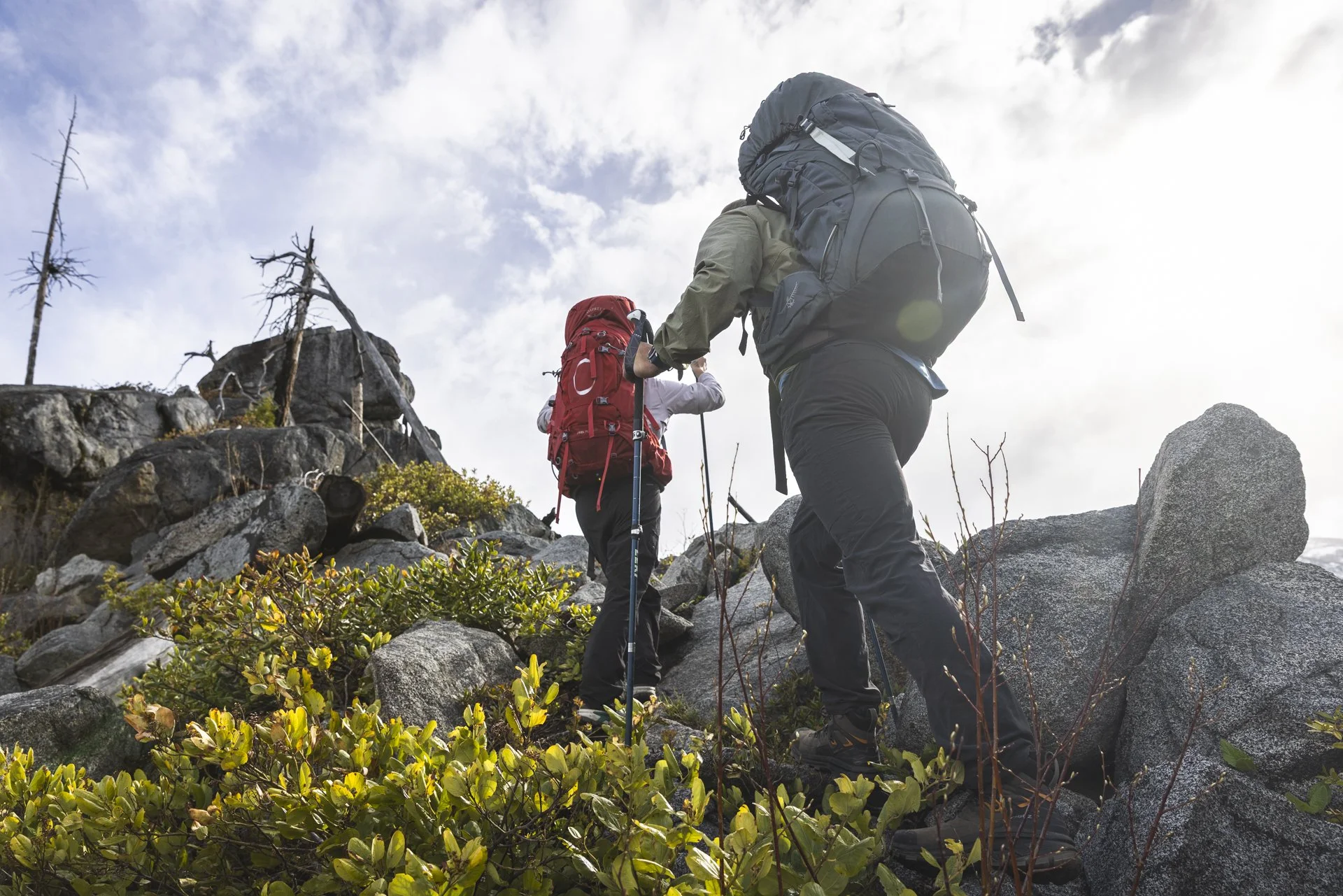 Two hikers ascending a rocky mountain trail with backpacks and trekking poles, surrounded by sparse vegetation and a cloudy sky.