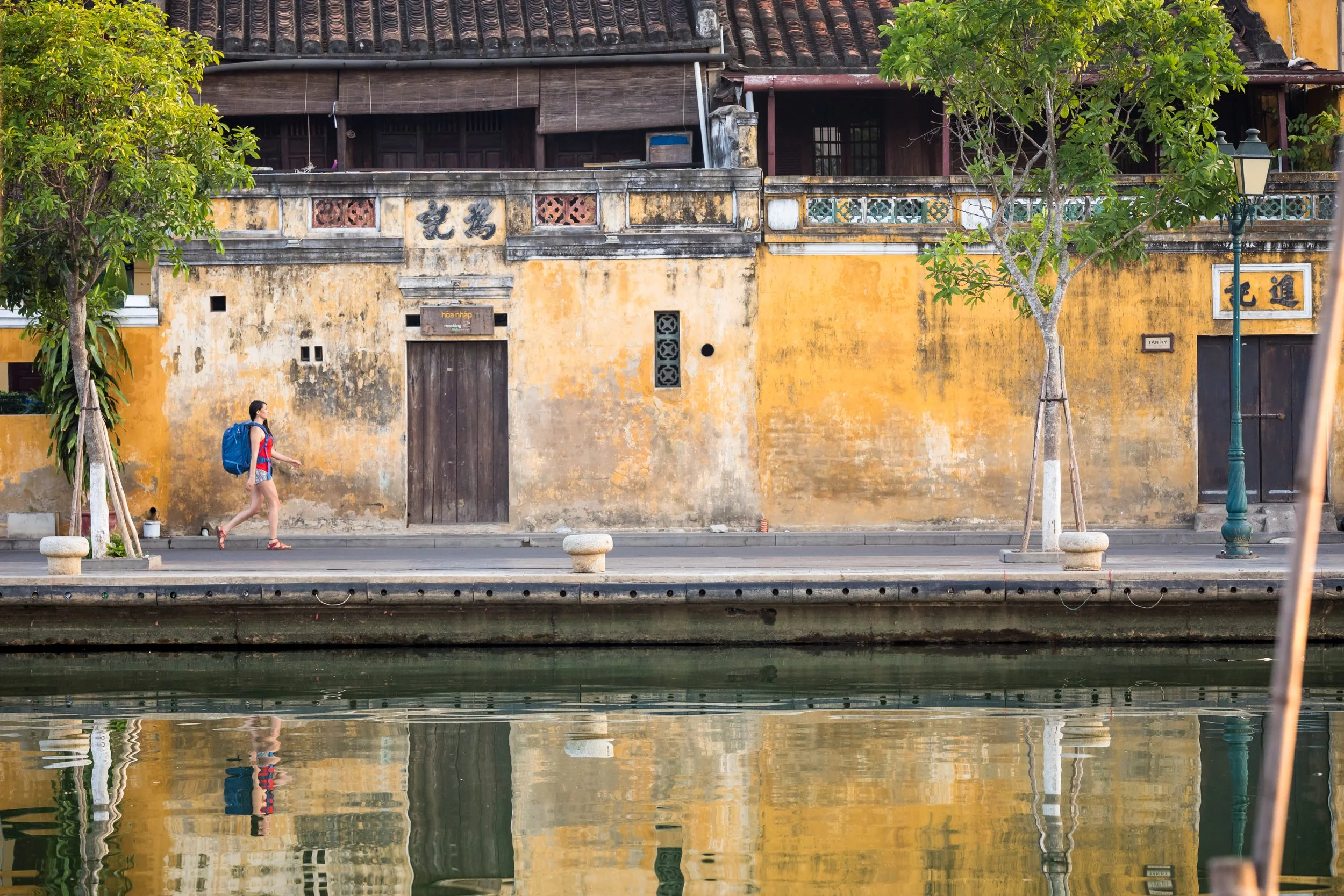 A woman with a blue backpack walking along a sidewalk next to a yellow, weathered wall with signs of aging, near a body of water. There are trees and a streetlamp along the sidewalk. Commercial lifestyle production by Fieldborn Creative.