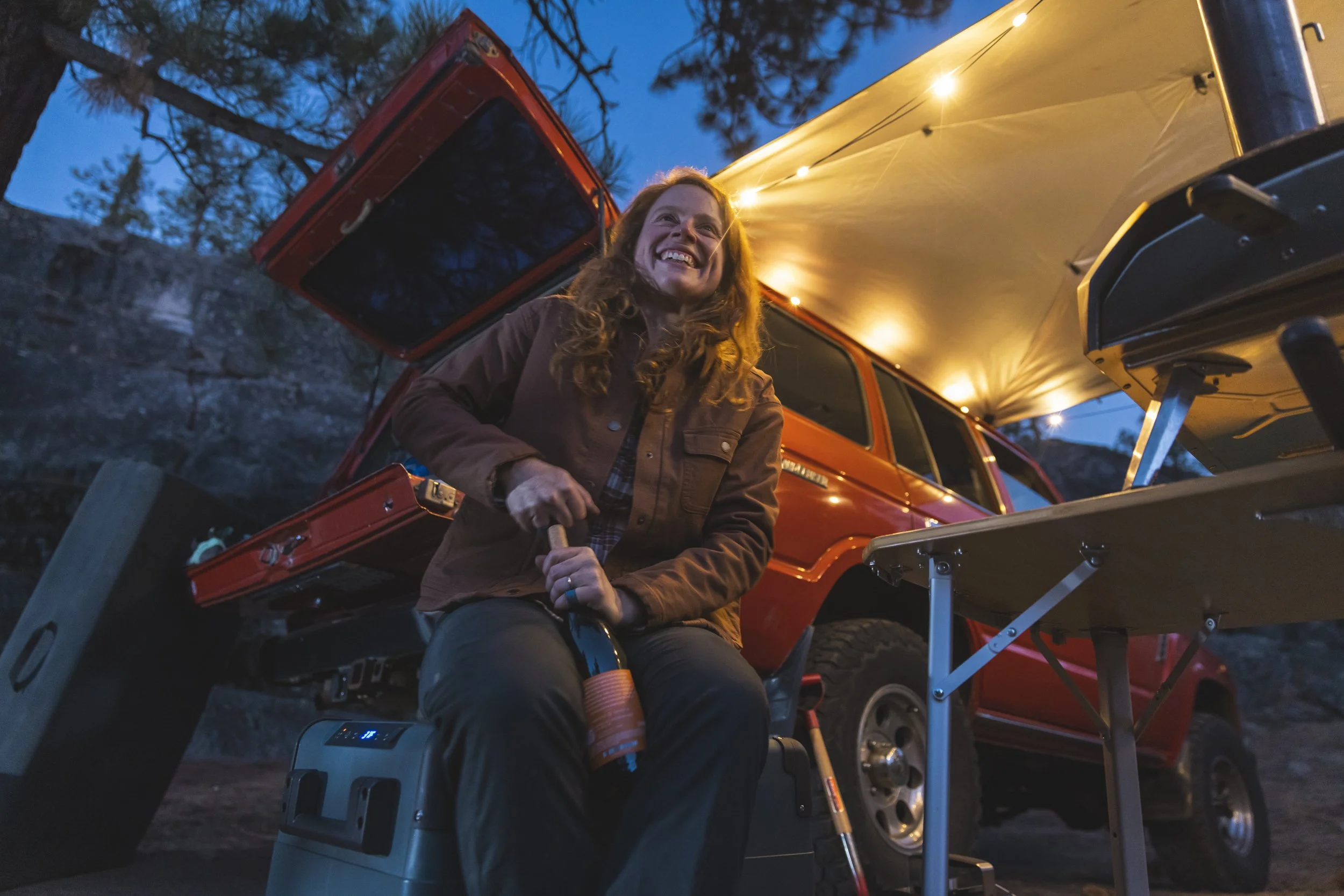 Red-headed woman opening a bottle of wine near a tailgate under an awning during a Dometic outdoor lifestyle shoot in California with string lights above, photographed by Fieldborn Creative.
