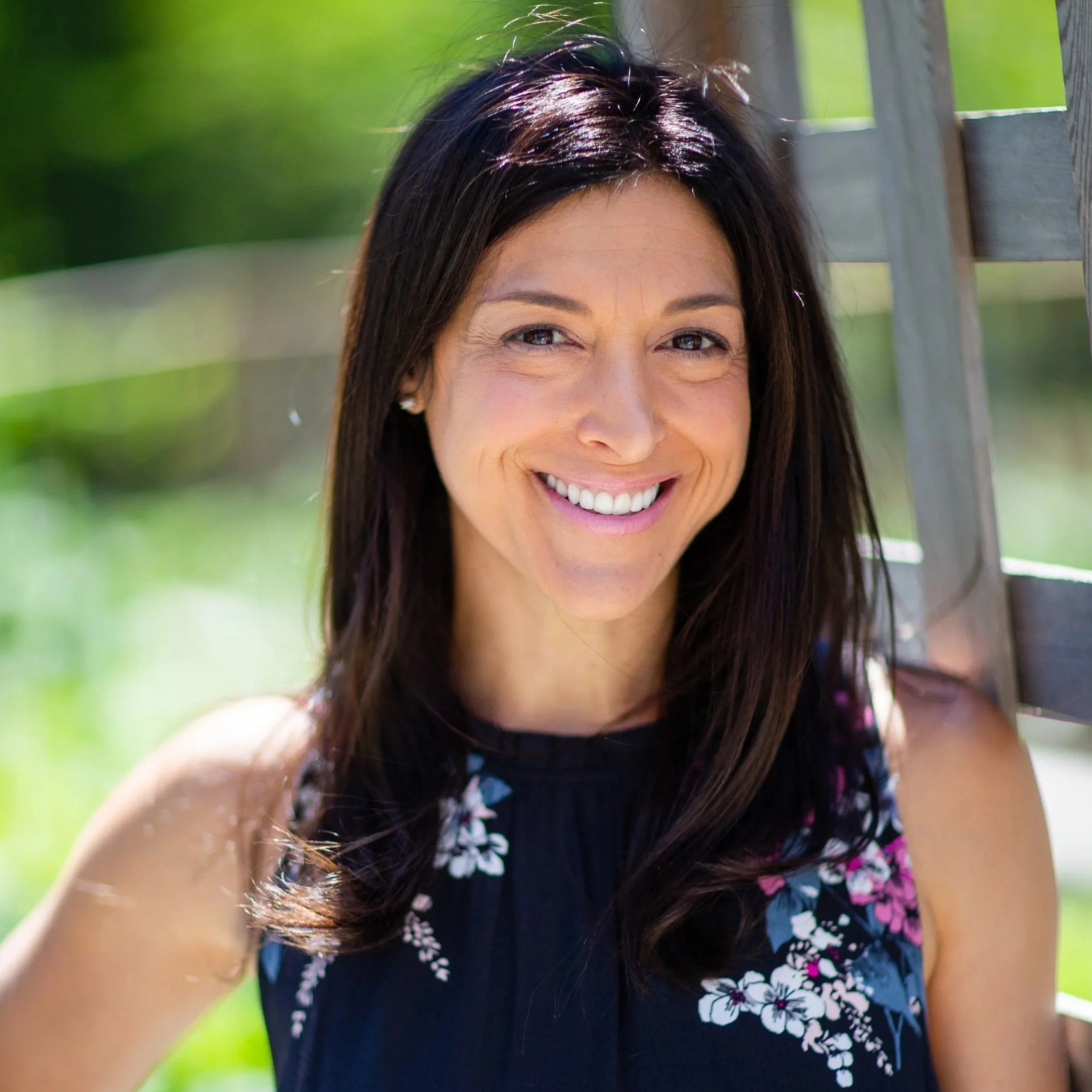 Smiling woman with dark brown hair wearing a sleeveless navy top with floral pattern, posing outdoors near a wooden fence.