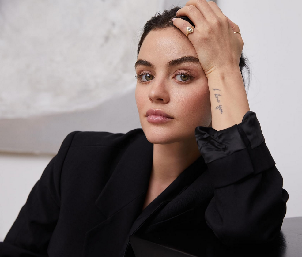 Actress Lucy Hale in a black blazer leaning on a counter with her hand on her head, in elegant jewelry and a tattoo that says ‘I love you’ on her outer wrist.