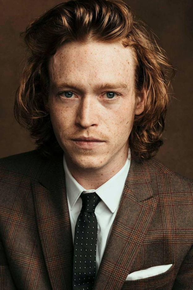 Actor and Musician Caleb Landry Jones in a brown suit and black tie. He has red hair, green eyes, and freckles.