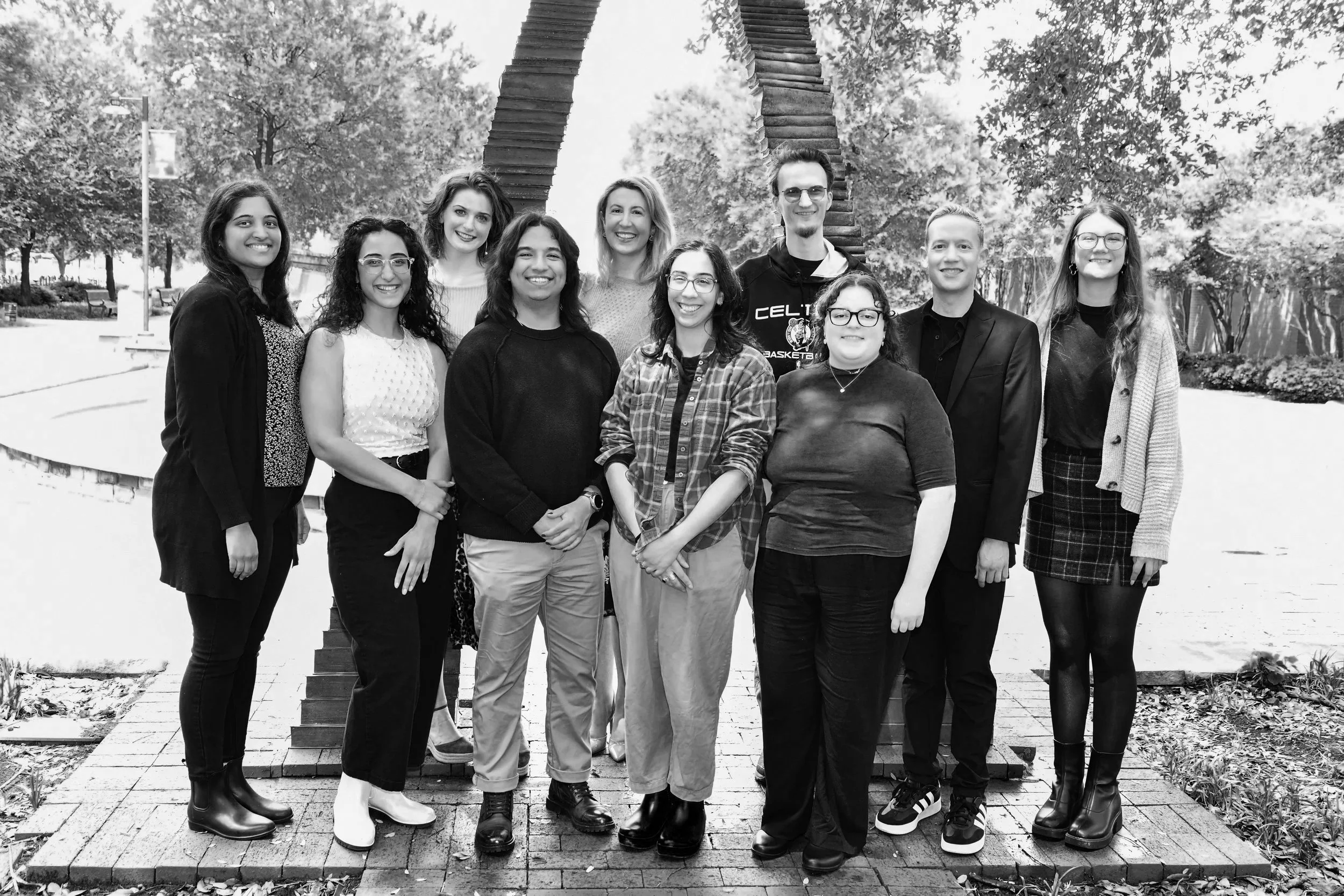 Group of eleven diverse young adults standing outdoors in front of a sculpture in a park, smiling at the camera.