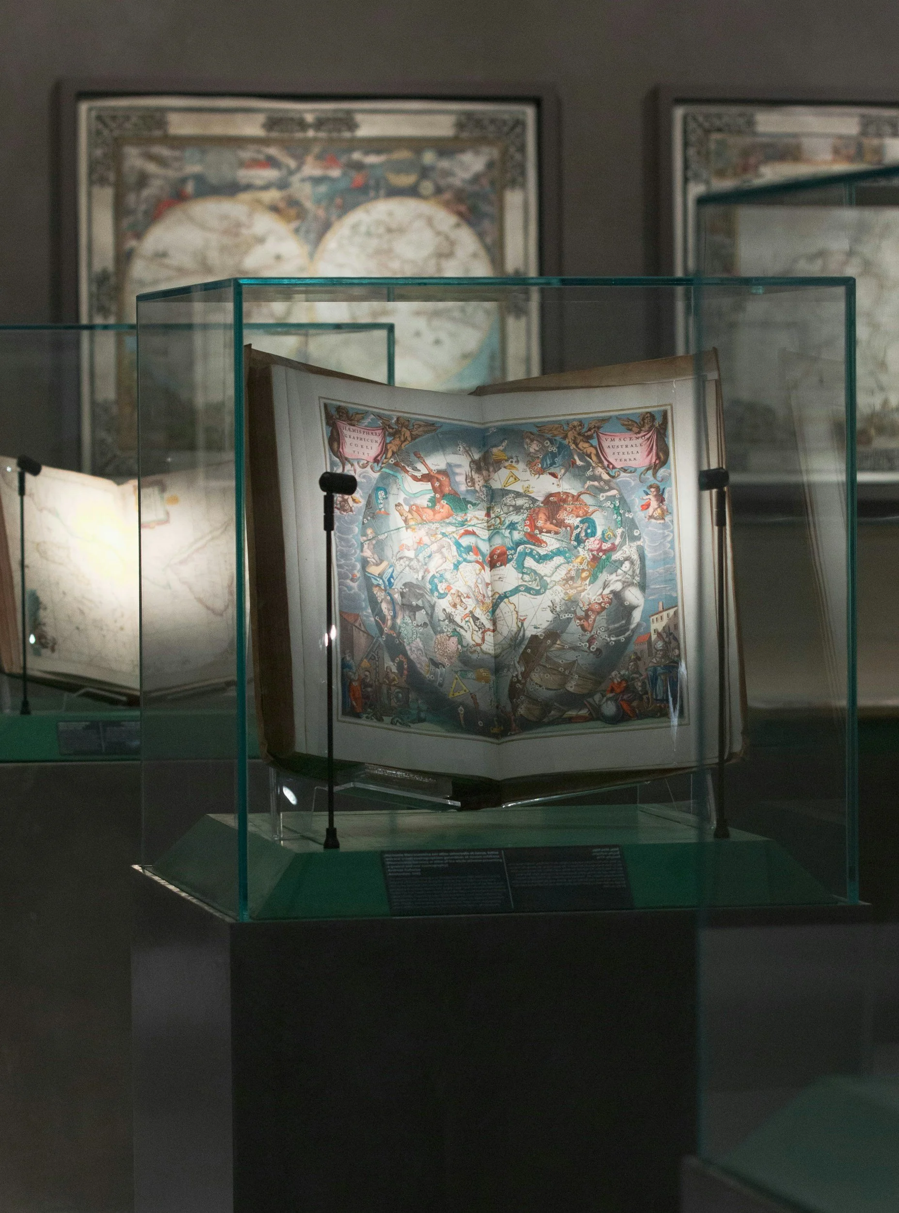 A display case holding an open, colorful illustrated book with medieval or mythological artwork, in a museum setting with framed maps or paintings in the background.