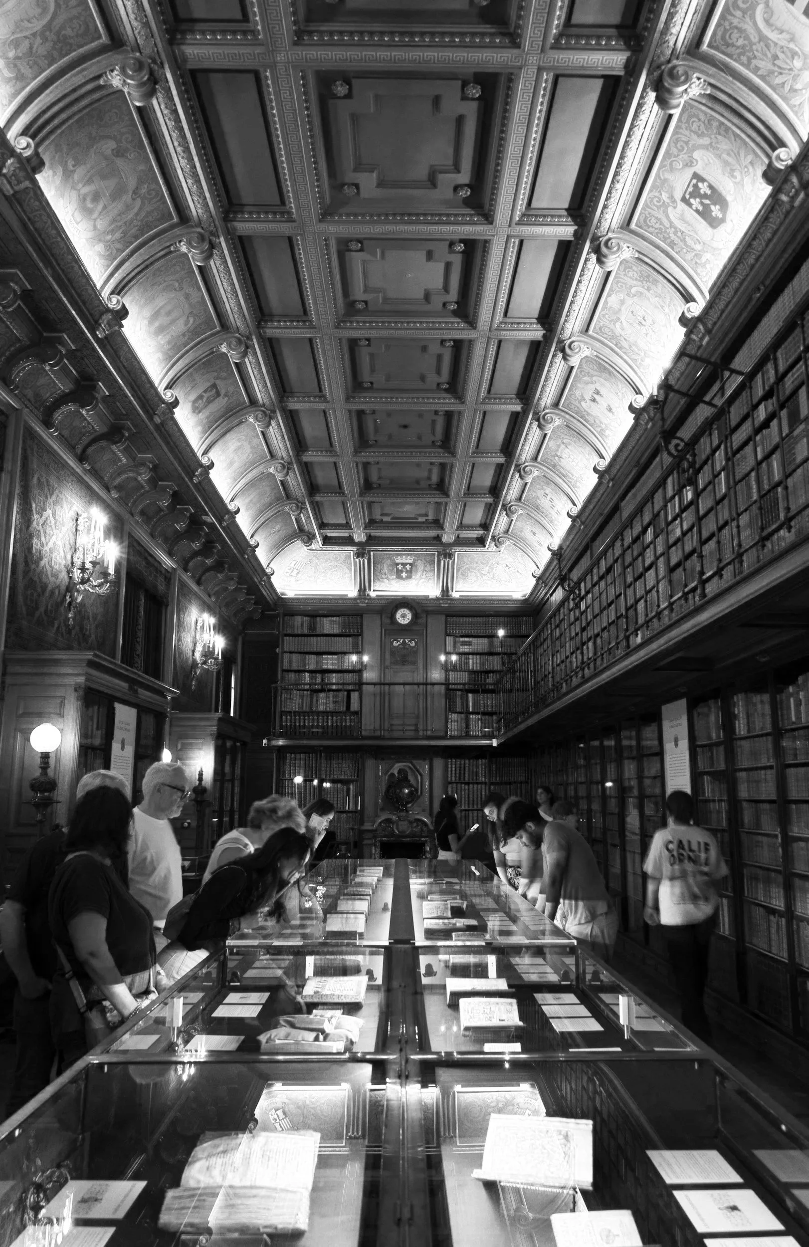 A black and white photo of a historic library with ornate ceiling, wood-paneled walls, and floor-to-ceiling bookshelves. Several people are observing displayed books through glass cases in the center of the room.