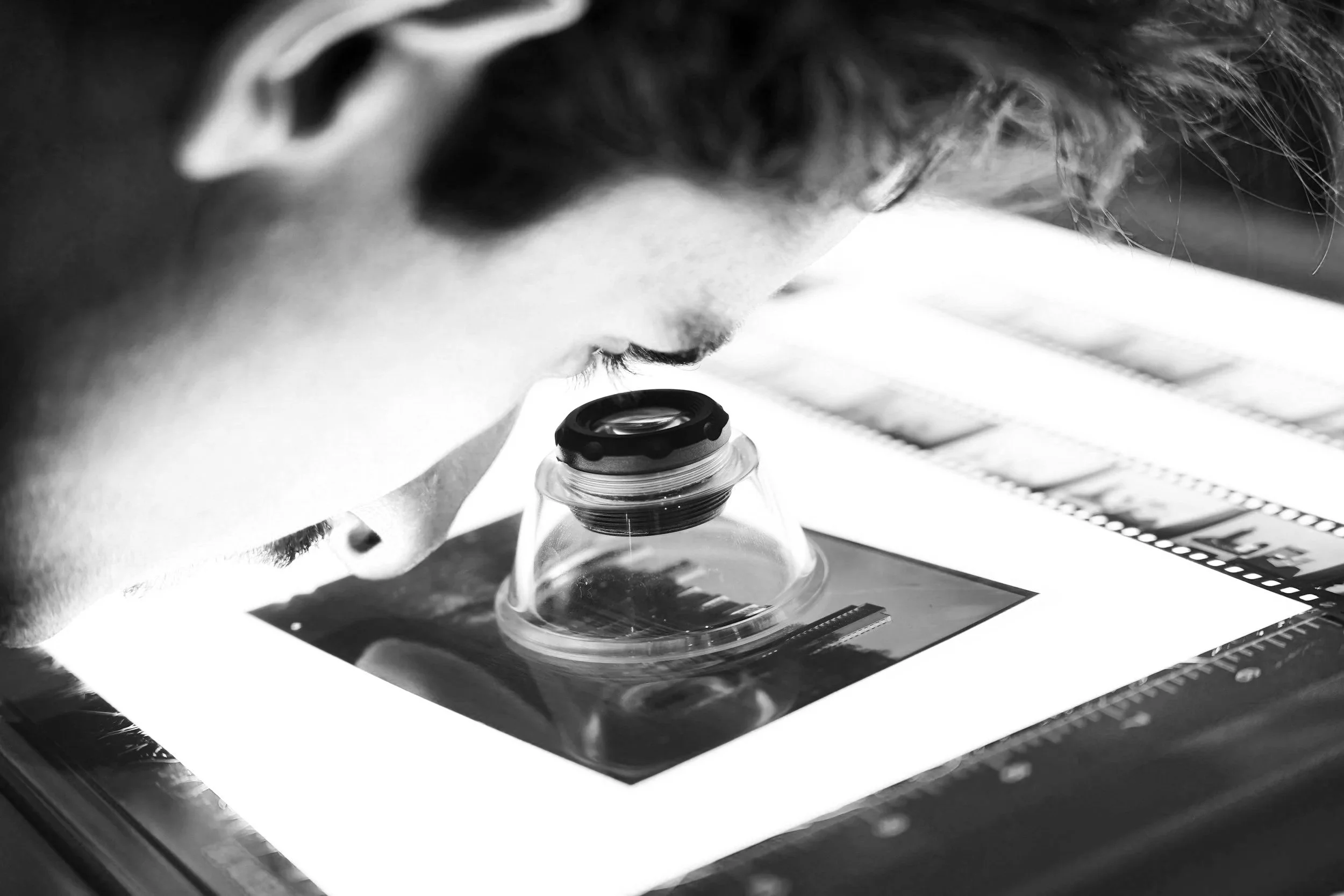A person looking through a microscope at a glass slide with film strips in the background.